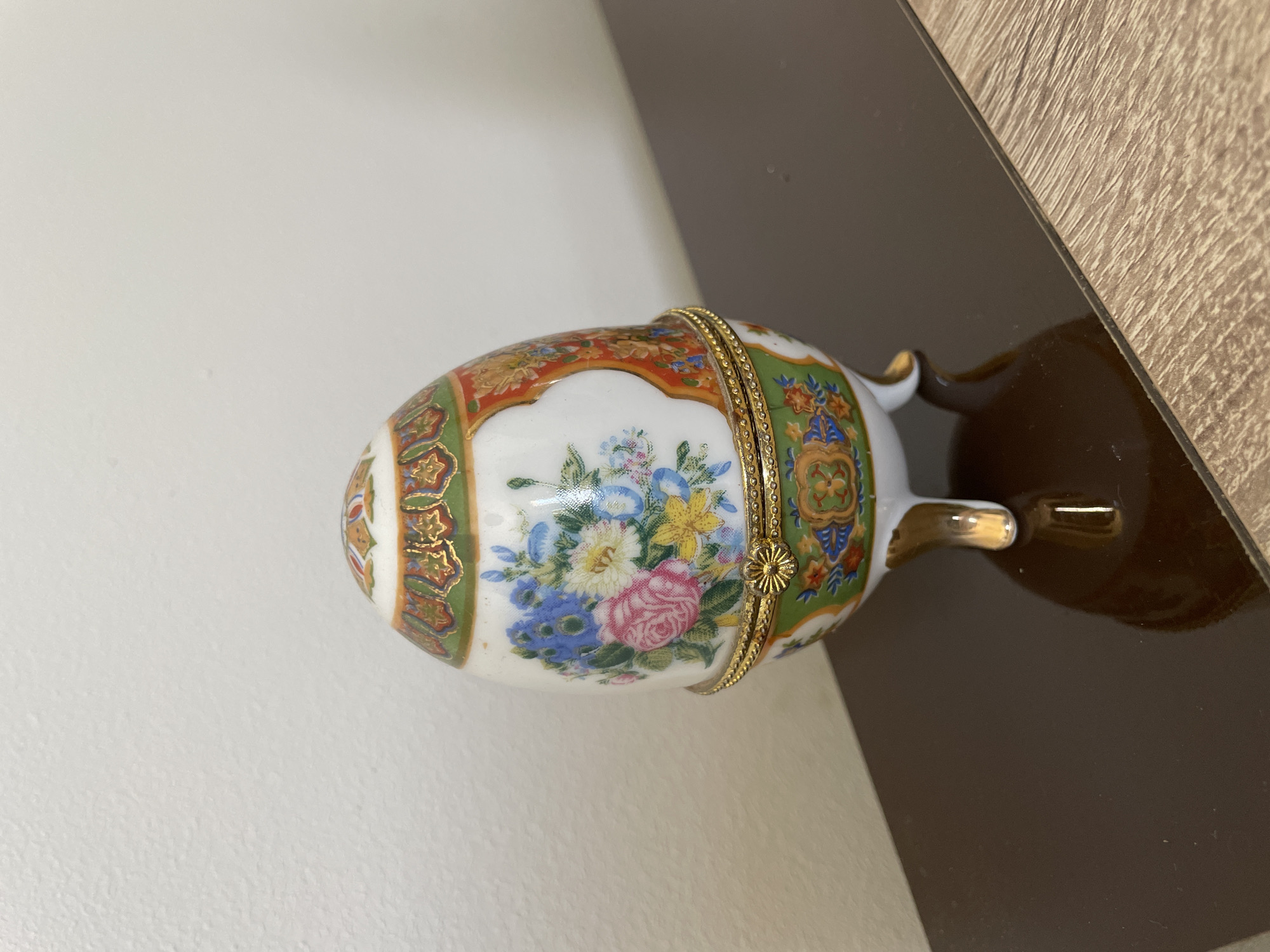 A small, ornate, egg-shaped porcelain box with a hinged lid sits on a dark, reflective surface. The box is decorated with a floral motif featuring roses, daisies, and other flowers in shades of pink, blue, yellow, and white, outlined in gold. The top of the box has a decorative band with a gold and red pattern. The bottom of the box has three small, gold-colored feet. The surface it rests on appears to be a shelf or table with a wood-grain texture. The background is a plain, textured white wall. The overall impression is one of delicate craftsmanship and decorative artistry. The scene is indoors.
