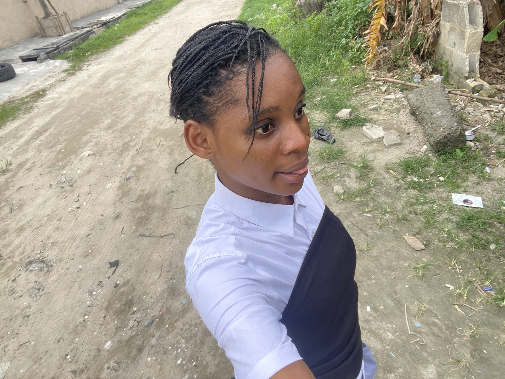 A medium-shot photograph captures a young woman from a slightly high-angle, looking off to the right. She has dark braided hair pulled back, and is wearing a white collared shirt with a black garment layered over it.
The background consists of a dirt and concrete path leading into a cluttered area of grass, concrete debris, and refuse. The setting is in Iranla, Nigeria. The condition of the path and surrounding environment suggests a semi-urban or possibly peri-urban area.