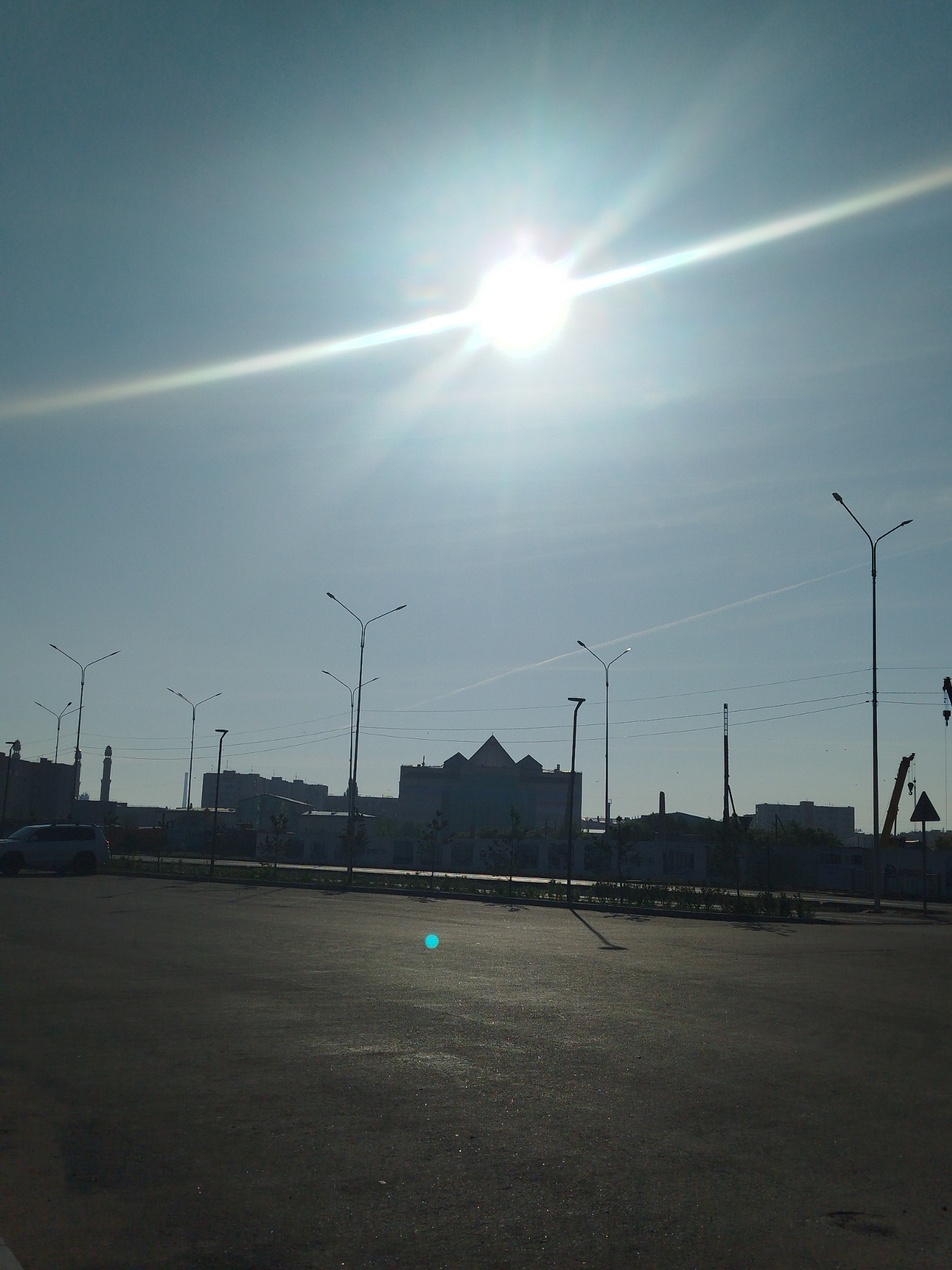 The image depicts an outdoor scene under a bright, slightly hazy sky with the sun intensely shining, creating a lens flare effect. In the foreground, a dark asphalt surface, likely a road or parking lot, is visible. A white SUV is parked on the left side of the image, facing away from the camera.
The middle ground shows a row of buildings, some appearing industrial or administrative, with a distinct minaret on the far left, suggesting a location in Kazakhstan, specifically Kokshetau, given the context provided. Several streetlights are positioned along the road. Construction cranes are visible in the background on the right, indicating ongoing development or industrial activity. The sky has a faint contrail. The overall impression is an urban or semi-urban environment on a sunny day.