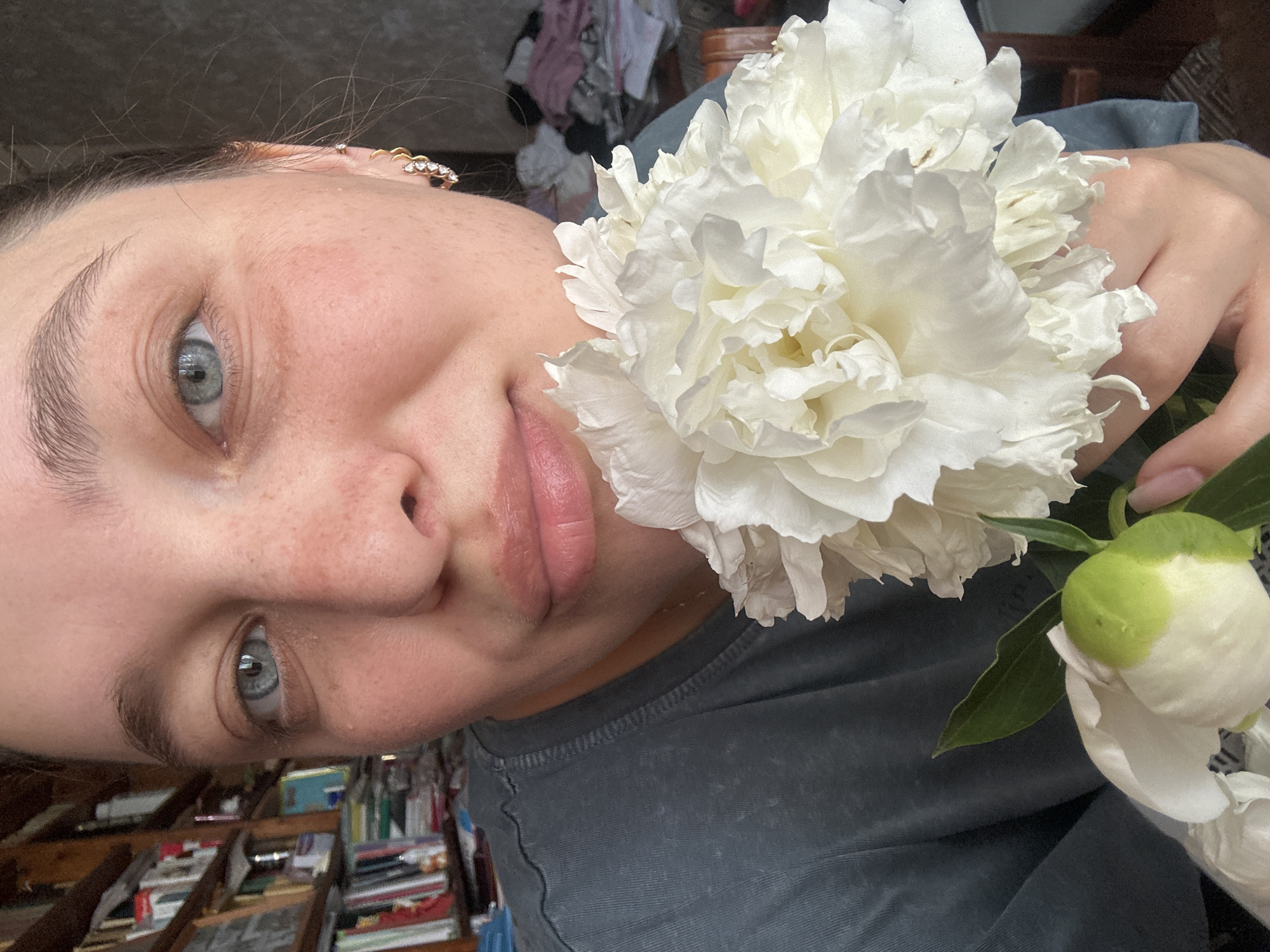 A close-up, eye-level, indoor, natural-light shot shows a young person with light blue eyes and fair skin with freckles holding a large, white peony. The person is looking towards the camera with parted lips. A small, green peony bud is also visible at the bottom left of the frame. The person is wearing a dark gray t-shirt. In the background, a bookshelf filled with books is on the left, and a blurry, cluttered area with clothing is on the right. The scene is likely indoors, possibly in a residential setting. No specific location in Yaroslavka, Russia is identifiable from the image.