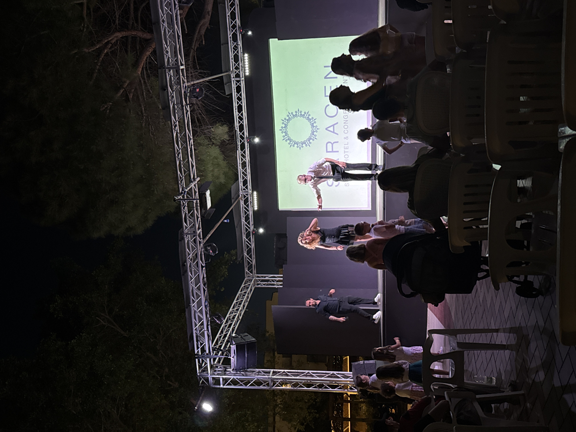 The image depicts an outdoor evening event at the Saracen Hotel & Congress Centre in Isola delle Femmine, Italy. On a raised stage, three performers are visible. To the left, a man in a black shirt and pants stands with his arms at his sides. In the center, a woman in a black dress with a short skirt is gesturing with her right hand. To the right, a man in a white shirt and dark pants with a tie is pointing his left arm forward. Behind the performers, a large screen displays a logo and the text "SARACEN HOTEL & CONGE". The stage is lit by spotlights. In front of the stage, an audience is seated on chairs and some individuals are standing. Among the audience, several children are visible, some standing and watching the performance, while others are seated or in a stroller. The ground is paved with patterned tiles. Trees are visible in the background, suggesting an outdoor setting, and the overall lighting indicates it is after sunset.