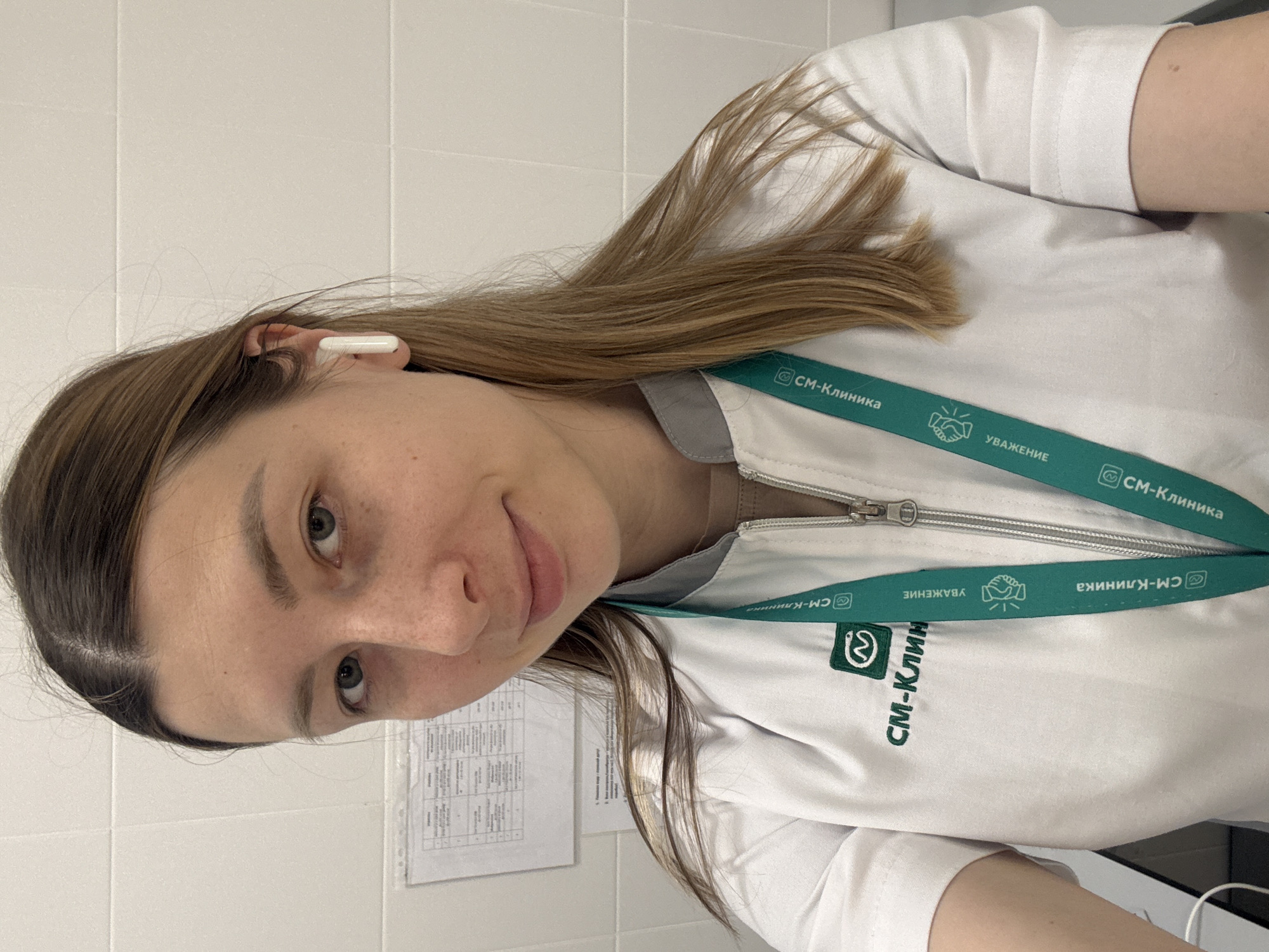 A young woman, wearing a white medical coat with a green lanyard that reads "CM-Клиника" (SM-Clinic) and "УВАЖЕНИЕ" (Respect), poses in what appears to be a medical setting. She has light brown hair, fair skin, and is wearing a white wireless earbud. A document is attached to the wall. The text on the lanyard and coat suggests this is likely taken at or near the CM-Clinic, a medical facility located in Moscow, Russia.