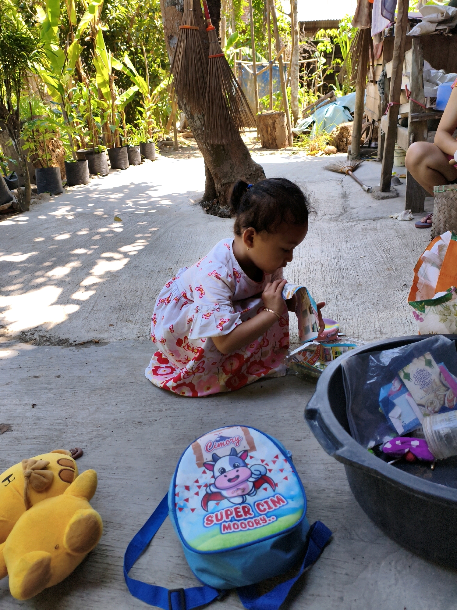 In Karangtalun, Indonesia, a young girl with dark hair tied in a ponytail is crouched on a concrete ground amidst tropical foliage. She wears a white dress with a red floral pattern. The girl is holding a colorful paper object up to her face, seemingly examining it. Beside her lies a blue backpack with a cow cartoon character and the words "SUPER CIM MOOOOORY" printed on it. Two yellow plush toys, possibly a giraffe and a bear, are on the ground to the left of the backpack. A large dark basin, partially filled with various items including plastic bags and colorful wrappers, sits to the right. In the background, several potted plants are arranged in a row, and two brooms are propped against a tree. The scene is bathed in sunlight, creating dappled shadows on the ground. A partial view of another person's legs and feet, wearing flip-flops, is visible on the far right.