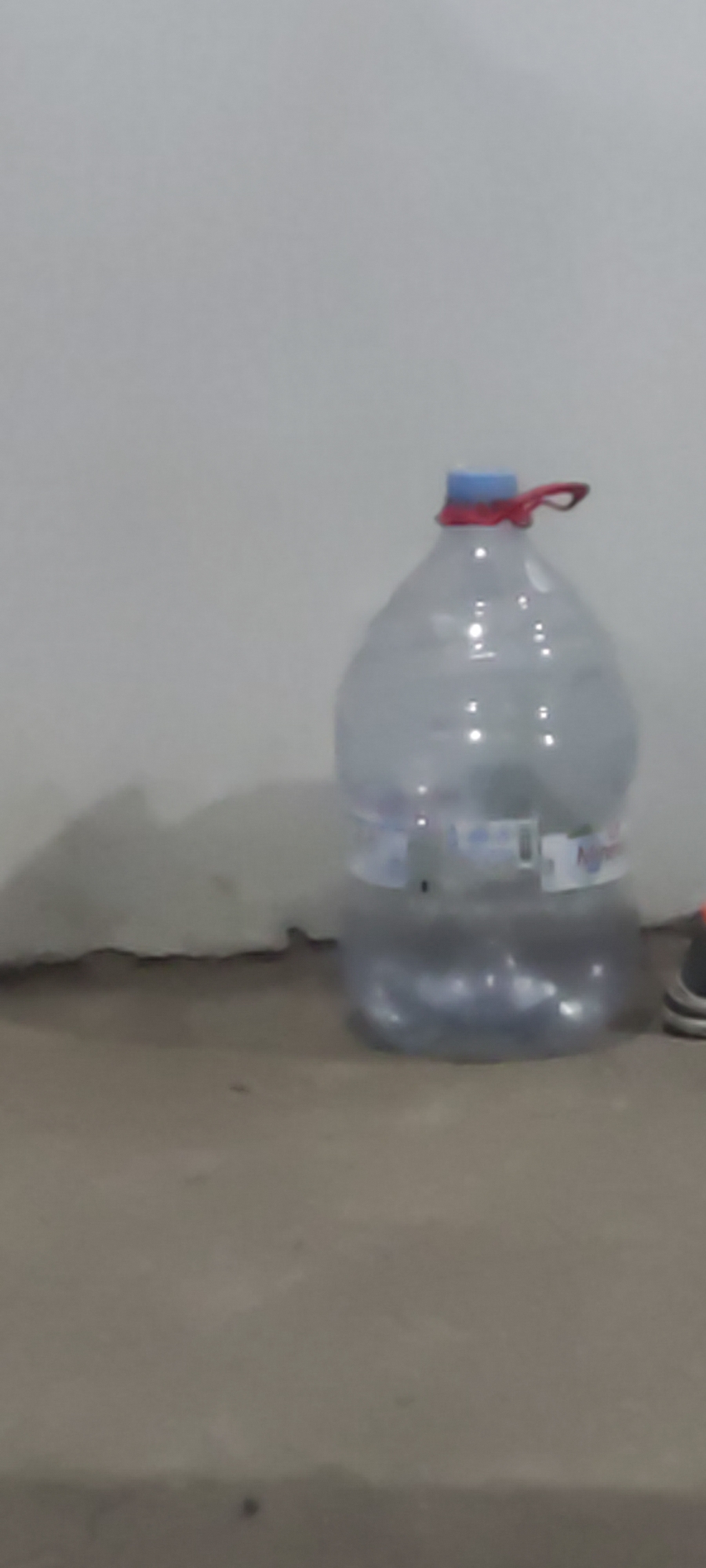A large, clear plastic water jug with a blue cap and a red loop handle is positioned on a concrete floor. The jug is partially filled with water and appears to be sitting against a light-colored wall with a dark, irregular line at the base of the wall, possibly indicating a baseboard or a shadow. A portion of a dark-colored shoe with an orange accent is visible on the right side of the frame, near the water jug. The environment suggests an indoor setting, possibly a storage area or a room in a residence, in Karangwangun, Indonesia.