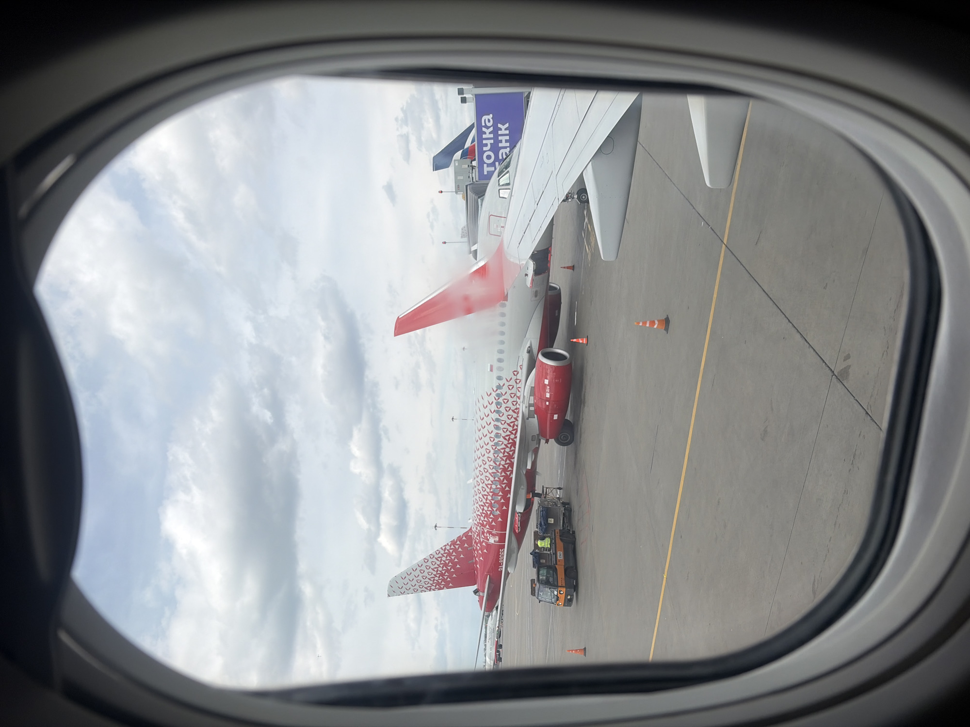 The image captures a view from an airplane window looking out onto an airport tarmac on a cloudy daytime. In the foreground, the gray frame of the airplane window partially encloses the scene, with a portion of the wing and engine nacelle of the viewing aircraft visible.
The primary subject is another airplane, parked on the tarmac. This aircraft is predominantly red and white, featuring a distinctive livery with a pattern of small, overlapping red and white geometric shapes that become denser towards the rear fuselage and tail fin. Its tail is red. A ground support vehicle, likely a baggage tug, is positioned near the rear fuselage of this red and white plane, with at least one ground crew member visible wearing a high-visibility vest. Several orange traffic cones are placed on the tarmac around the parked aircraft.
In the background, a large blue sign with white Cyrillic text "ТОЧКА БАНК" (Tochka Bank) is visible, suggesting it's mounted on a jet bridge or an airport building. The sky above is largely overcast with patches of lighter clouds. The tarmac itself is gray with yellow painted lines.
The scene depicts typical airport ground operations, likely preparing for departure or post-arrival procedures. Given the "Точка Банк" text and the provided context of Saint Petersburg, Russia, the setting is an airport in Russia. The red and white patterned livery is characteristic of certain Russian airlines, such as Azimuth Airlines.
