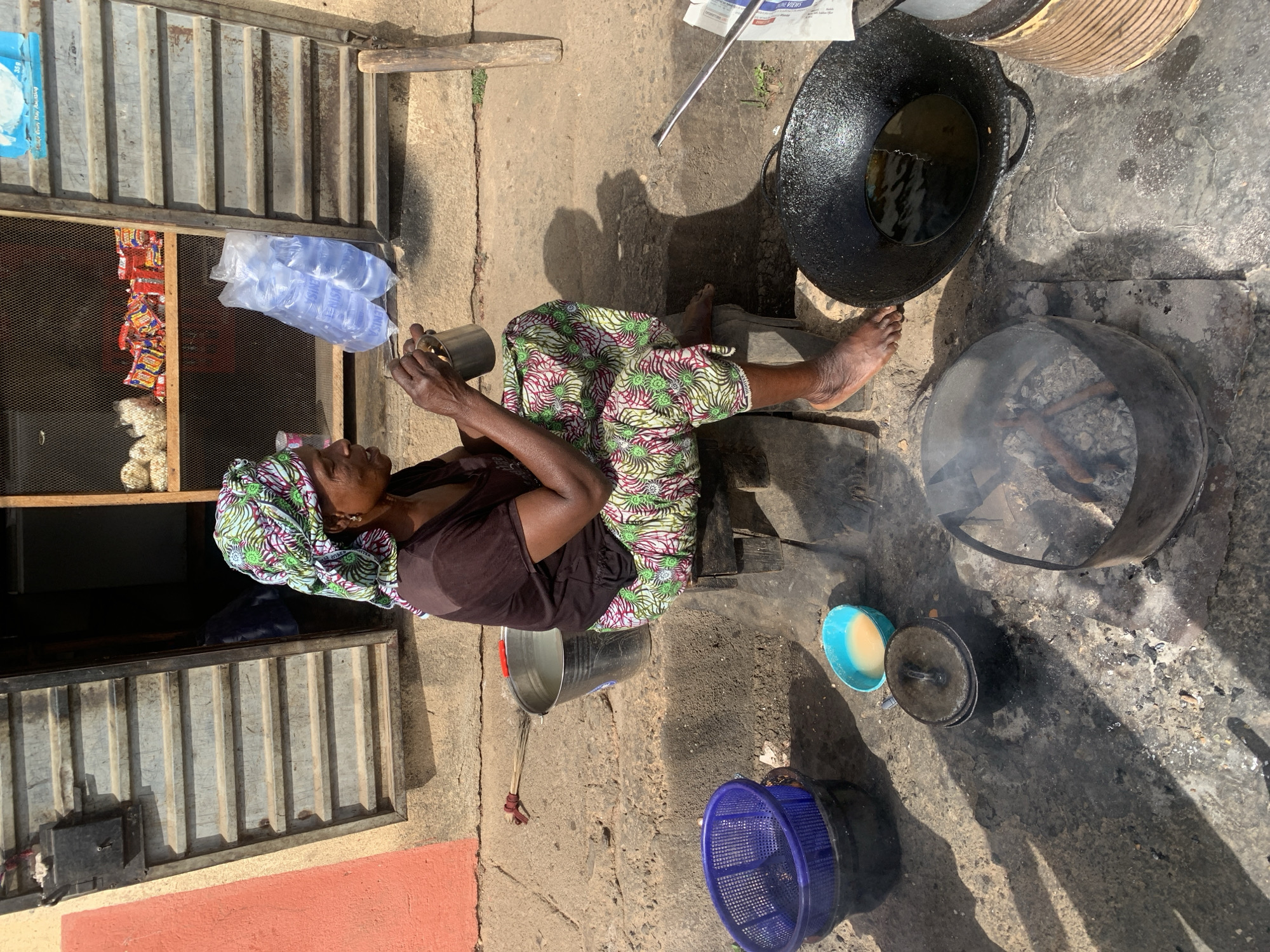 This image captures an outdoor scene in Jos, Nigeria, depicting a woman engaged in what appears to be food preparation or selling activities. The setting suggests a small, informal street food stall or shop.
A woman, wearing a vibrant green, red, and white patterned skirt or trousers and a dark brown top with a patterned headwrap, is seated on a low wooden stool. Her bare feet are visible on the concrete ground. She holds a small metal cup in her hands, her gaze directed downwards, seemingly focused on an immediate task.
Around her, various cooking and storage items are arranged. To her right, a large, black cast-iron wok filled with oil sits on the ground, suggesting deep-frying is underway or about to begin. Adjacent to it, an open fire burns inside a round metal brazier, producing a visible plume of smoke, indicative of cooking with charcoal or firewood. A smaller lidded pot and a light blue bowl containing a yellowish liquid are also on the ground nearby. Further back, a blue plastic basin and a large metal bucket are present.
In the background, a structure with corrugated metal panels likely serves as a shopfront or storage area. Inside, shelves display packets of various snacks, and two clear plastic bags containing multiple sachets of drinking water, labeled with "BLUE" and possibly "PURE WATER," hang prominently. A piece of newspaper is visible near the large wok.
The scene is illuminated by bright sunlight, casting distinct shadows across the concrete ground, suggesting it is daytime. The overall atmosphere conveys a sense of daily life and commerce in a bustling, outdoor environment. The woman appears calm and focused on her work.