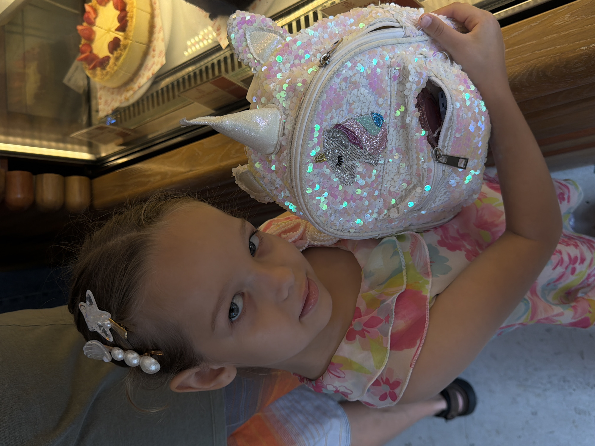 A girl with fair skin and blue eyes is looking up at the camera. She has brown hair with two barrettes: one with a star and one with pearls. She is wearing a pink and white floral dress. A white, sequined unicorn backpack is partially covering her. Her right hand is holding the backpack open, revealing an unknown object inside. A display case with cakes is in the background. The location is identified as Bac Nha Trang Ward, Vietnam.