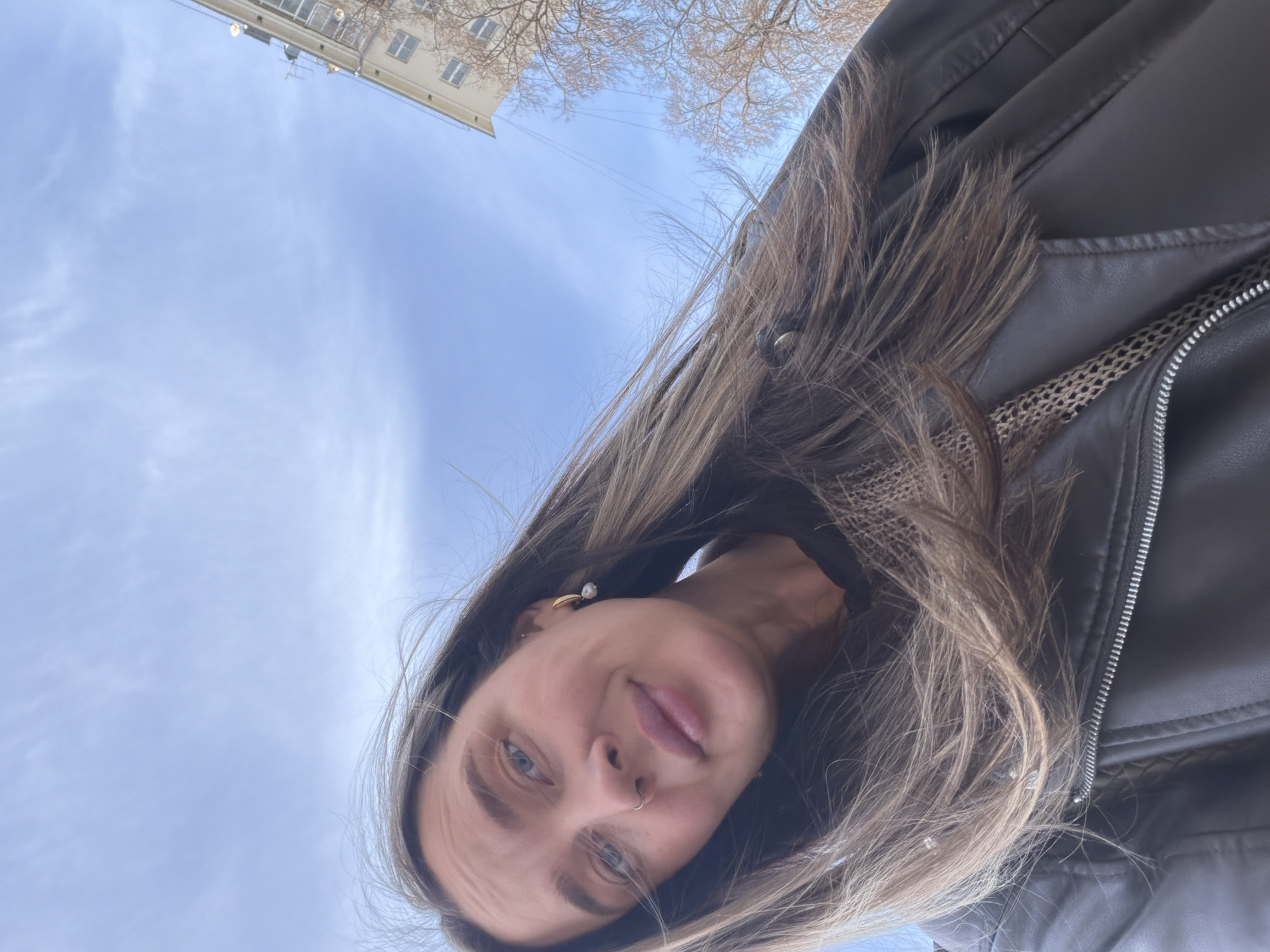 The image shows a person with light brown hair and blue eyes. They have a nose ring and an earring with a pearl. They are wearing a black leather jacket. The background shows a blue sky and a building. The building appears to be located in Moscow, Russia, based on the architectural style, specifically of residential type. The photograph is taken from a low angle, looking up at the person and the sky.