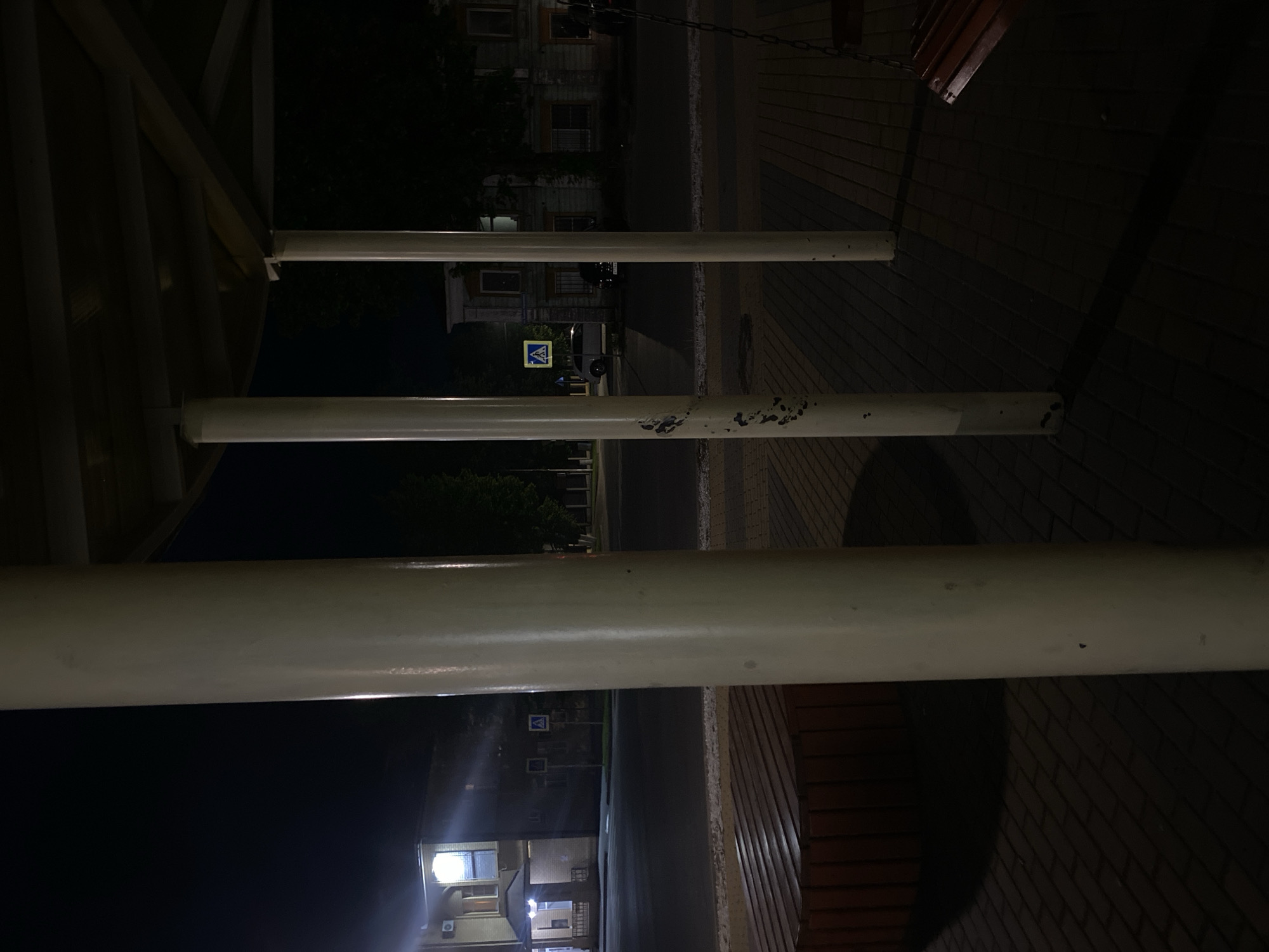 The media file captures a nocturnal outdoor scene from a low vantage point, possibly from beneath a covered structure. Three prominent, horizontal white cylindrical poles dominate the foreground, extending across the frame and partially obscuring the background. The poles show some minor scuffs and dark marks, suggesting wear.
To the left, a darker, multi-paneled structure, likely part of a roof or shelter, is visible. To the right, a textured dark surface, possibly brick paving or a wall, recedes into the distance.
In the mid-ground, a street or pathway is visible. A clear yellow triangular sign with a blue border and a blue symbol of a person crossing (a pedestrian crossing sign) stands out against the darkness. A light-colored vehicle is partially visible near the sign. Further in the background, several buildings with illuminated windows can be seen, hinting at distant artificial light sources. Sparse trees are also discernible against the dark night sky. No individuals are present in the image. The scene is lit by artificial lights from buildings and possibly streetlights, creating deep shadows and highlights on the poles. The overall impression is quiet and still, characteristic of a late-night urban or suburban environment.