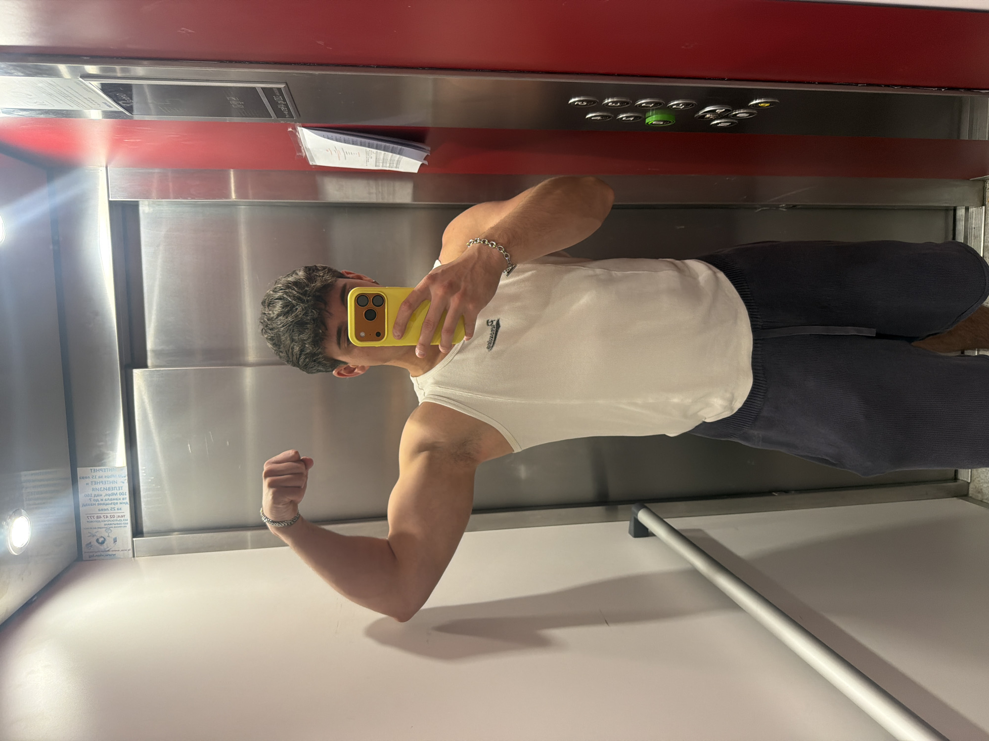 A young man is captured in a mirror selfie inside an elevator, flexing his left bicep. He has short, dark, curly hair and is wearing a white ribbed tank top and dark trousers. His face is mostly obscured by a bright yellow smartphone with a brown camera case, which he holds up to take the photo. A silver chain bracelet adorns his left wrist.
The elevator interior features a combination of metallic, likely stainless steel, panels and red horizontal strips. A control panel with several silver buttons, one illuminated green, is visible on the right wall. A silver handrail runs along the lower portion of the wall. A bright light fixture is visible on the left wall, casting a glare.
A sign on the left wall displays text in Cyrillic, which, when deciphered, indicates "ИНТЕРНЕТ" (Internet) and mentions details such as "100 MB" and a date of "21 септември 2021" (21 September 2021), suggesting free Wi-Fi service. This Cyrillic text and details like a potential ".bg" domain in smaller print, along with the provided location context, confirms the setting is in Sofia, Bulgaria. The image depicts a casual moment in a modern building.