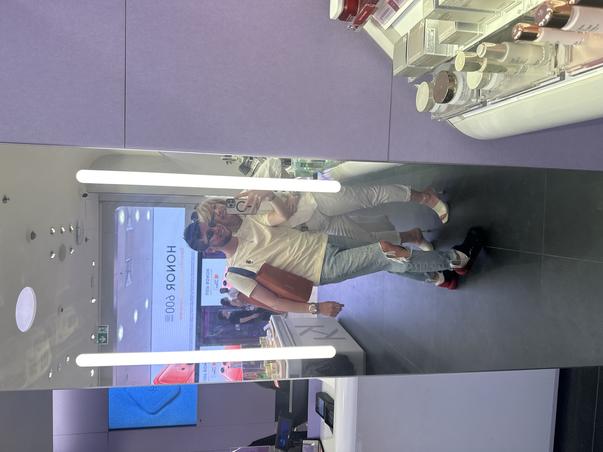 The image is a mirror selfie taken in a retail environment, likely a department store or a beauty store, in Moscow, Russia. A man and a woman are posing for the photo. The man, on the left, is wearing sunglasses, a light yellow t-shirt, ripped blue jeans, and black chunky shoes with red accents. He is carrying a large tan tote bag over his shoulder. The woman, on the right, is wearing a light-colored top and matching pants and white heeled sandals. She is holding a smartphone, taking the selfie. They are standing in front of a large mirror that reflects the store's interior. The background shows displays of products, possibly cosmetics, with shelves stocked with various jars and bottles. There are also digital screens visible in the reflection. The store has a clean, modern aesthetic with lighting fixtures integrated into the mirror and ceiling. The overall atmosphere suggests a shopping trip in a well-lit commercial space.