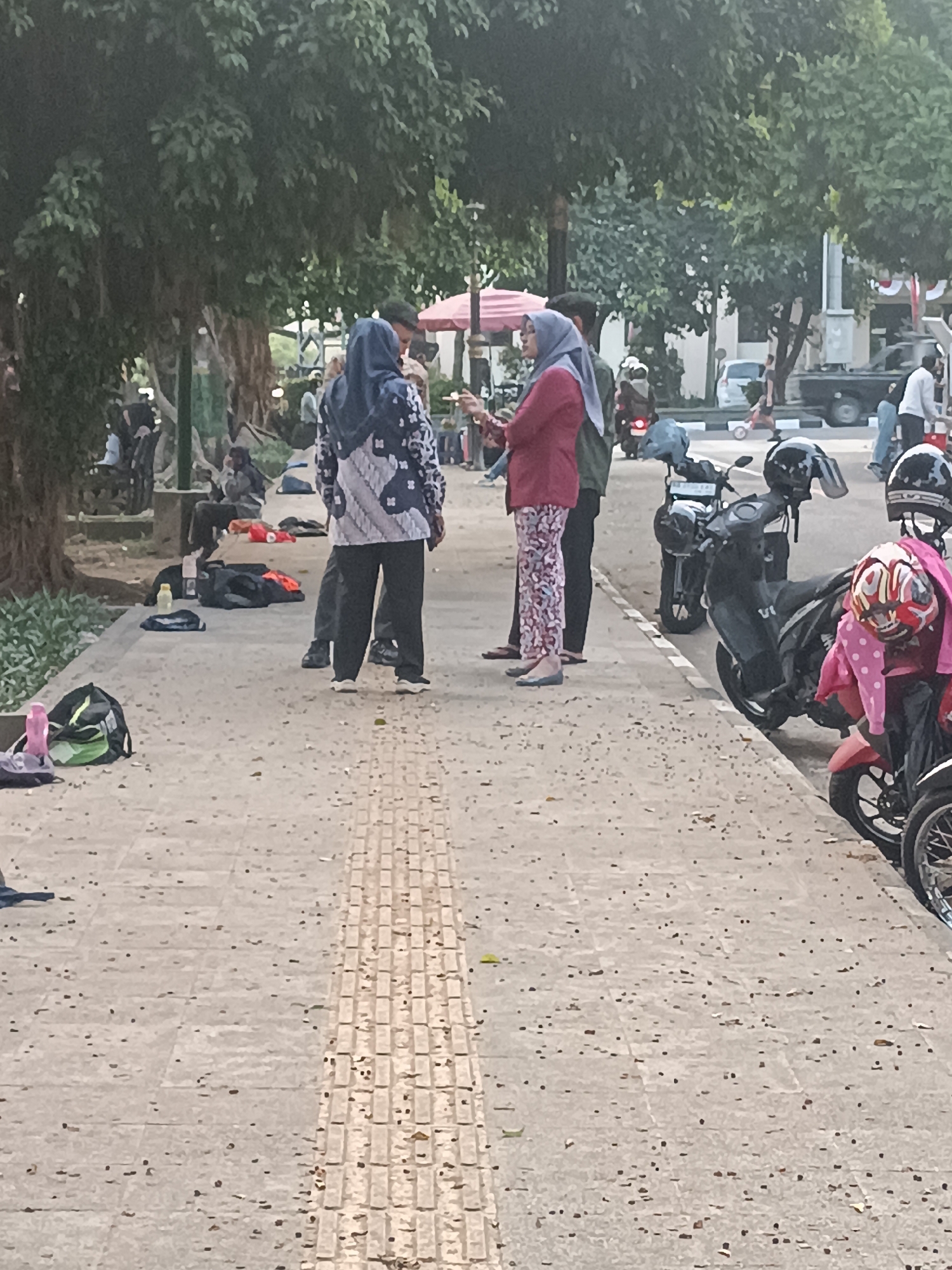 In Sleman, Indonesia, on a paved sidewalk with a tactile paving strip, several people are gathered near parked motorcycles. Two women, one in a dark blue hijab and a patterned top, and another in a light gray hijab and a red jacket, are conversing. Behind them, a man in a gray shirt and dark pants is partially visible. Several black and gray motorcycles are parked on the right side of the frame. A green backpack and a pink water bottle are placed on the ground in the foreground. Trees with dense foliage line the left side of the sidewalk, and in the background, more trees, a building, and a parked truck are visible. The scene appears to be an outdoor public space.