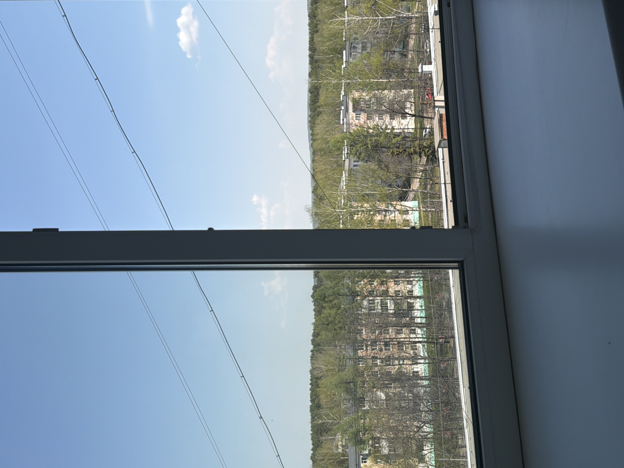 The image shows a view from a window, likely in an apartment building in Chaikovsky, Russia. Outside the window, there are buildings and trees with early spring foliage. Power lines are visible against a blue sky with a few scattered clouds. The window frame is white, and part of an interior wall is visible to the right. The scene depicts a typical urban landscape in a residential area during the daytime.