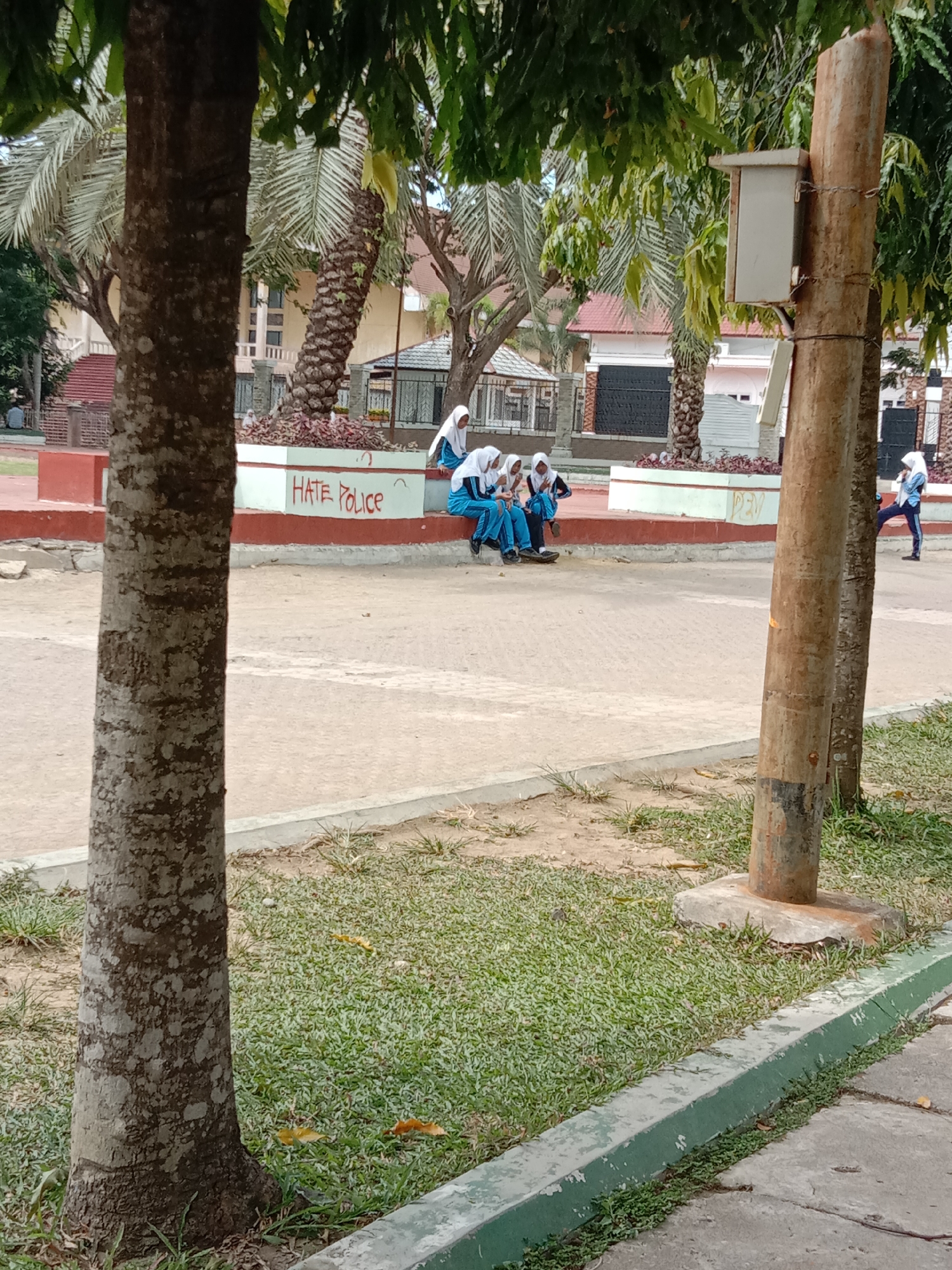 The image captures an outdoor scene in what appears to be a public space or courtyard in Lhokseumawe, Indonesia, under bright daylight. Two prominent tree trunks in the foreground, one on the left and one on the right, frame the view. A small, light-colored utility box is attached to the tree trunk on the right.
In the mid-ground, several young women or girls are visible, all wearing matching blue and white school uniforms along with white hijabs. Four of them are seated on a low, white concrete wall, which also serves as a planter box for reddish-brown plants. They appear to be resting or engaged in conversation. On the white wall, written in red graffiti, are the words "HATE Police." Further to the right on the wall, there are other faint, illegible markings. To the far right, another girl in a similar uniform stands with one leg raised, possibly stretching or in mid-stride.
The ground consists of a paved area in the center, flanked by patches of green grass, especially visible in the lower right corner where a green-painted concrete curb is present. In the background, numerous palm trees and other foliage are visible, along with various buildings, including some with reddish-brown roofs. The scene conveys a relaxed atmosphere on a sunny day.