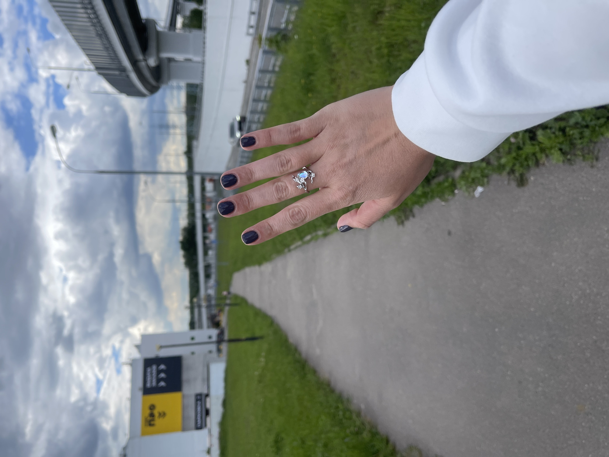A hand with a silver ring on the ring finger, featuring a light blue stone and floral accents, is presented against an outdoor backdrop in what appears to be Razvilka, Russia. The hand is wearing dark-colored nail polish and is attached to a white garment, possibly a sweater or jacket. Behind the hand, a gray paved pathway stretches into the distance, flanked by green grassy areas. A modern building with large signage is visible on the left side of the frame. Above, a cloudy sky fills the upper portion of the image. A highway overpass is present on the top left of the image.