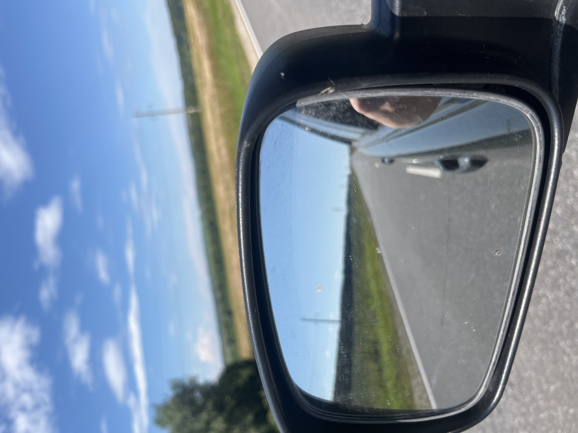 A close-up, slightly angled shot shows a car's side-view mirror, reflecting a bright blue sky with scattered white clouds. The mirror itself is rectangular and framed in black plastic. It is dirty with several smudges and small specks.
The reflection in the mirror reveals a rural landscape. A paved road stretches into the distance, flanked by green fields and some trees. A telephone pole is visible in the background, and a power line can be seen running across the frame. In the lower right portion of the mirror's reflection, a person's hand is visible.
The exterior of the car is partially visible outside the mirror. The environment appears to be outdoors on a sunny day. The image was taken from inside the vehicle, looking out. The scene does not provide specific information about the city of Kukshumy, Russia.
