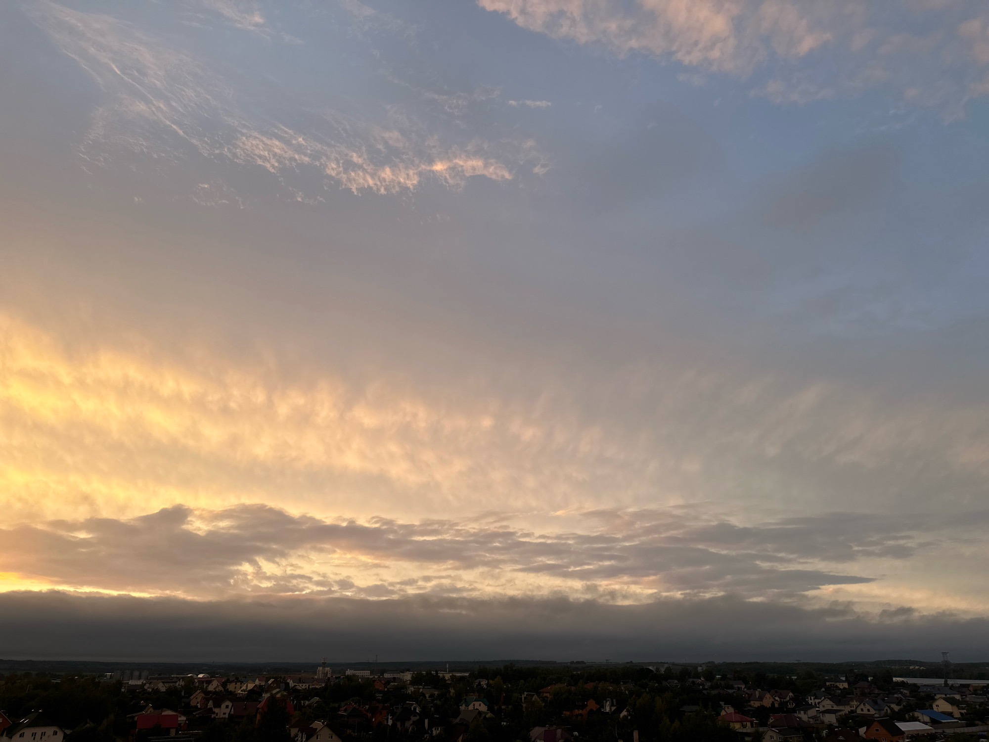 The image captures a panoramic view of a suburban landscape at dusk, with the sky dominating the upper two-thirds of the frame. A dense layer of clouds stretches horizontally across the middle ground, transitioning from a bright, golden hue on the left, illuminated by the setting sun, to a muted grey on the right. Higher in the sky, wispy cirrus clouds are visible against a backdrop of pale blue and soft pink. Below the clouds, a sprawling residential area is discernible, characterized by numerous houses with red and brown roofs, interspersed with trees and darker shadows. The silhouettes of buildings and structures are faintly visible against the twilight sky, suggesting a rural or semi-urban setting within the Шчомысліцкі сельскі Савет region of Belarus. No people or discernible actions are present in the scene.