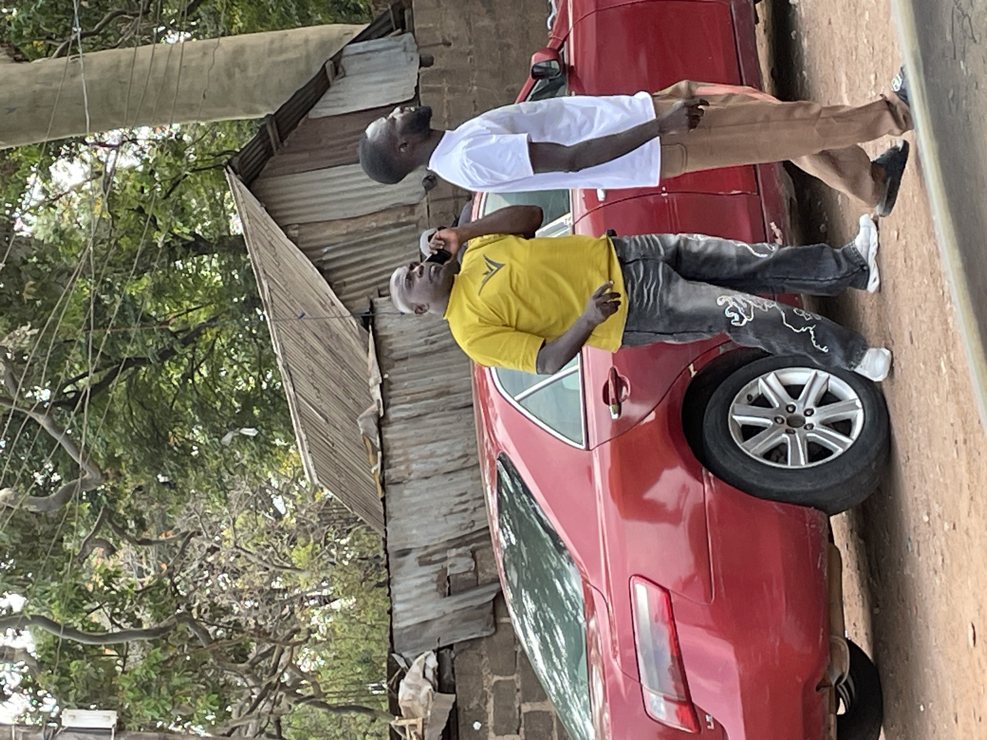 The image shows a street scene in Barakin Lerawi, Nigeria. Two men stand next to a red car. The man closest to the camera wears a yellow shirt and gray jeans with white embellishments and is talking on a cell phone. The other man wears a white shirt and khaki pants. A corrugated metal structure stands to the left of the car. There are trees visible in the background.