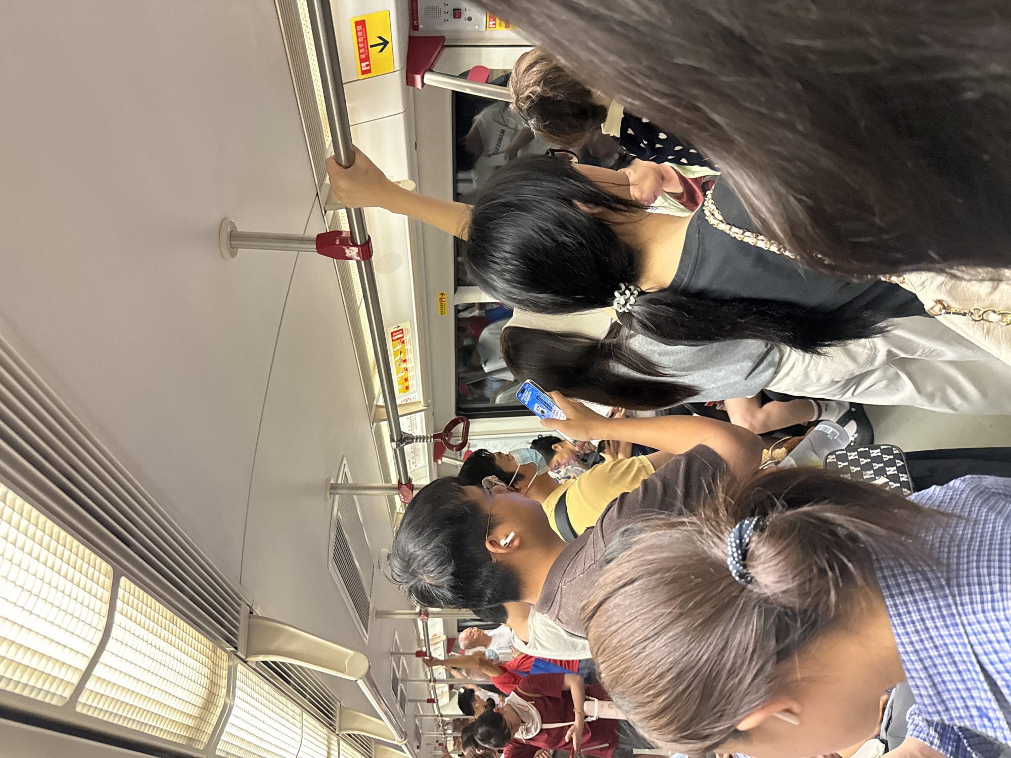 The image shows a crowded interior of a subway car, likely in Panyu District, China, during what appears to be peak hours. Passengers are standing and holding onto overhead handrails. The lighting in the car is fluorescent, with visible ceiling panels and ventilation grates. Several individuals are looking at their phones or wearing earbuds. One person in the foreground is holding a smartphone. A yellow sign with Chinese characters and a downward arrow is visible on the right side of the car, indicating an emergency exit or information. The overall scene is one of a busy urban commute.