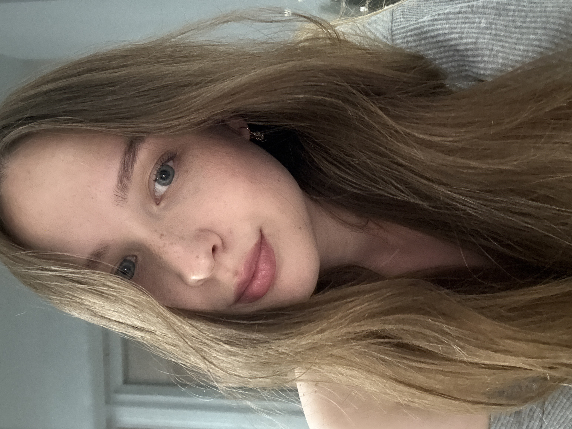 A close-up, head and shoulders selfie of a young Caucasian woman with long, wavy, light brown hair framing her face. She has fair skin with light freckles across her nose and cheeks. Her eyes are blue-grey, and she has light brown eyebrows. Her lips are slightly parted and appear to be wearing a light pink lip gloss. She is wearing a grey, striped shirt. A small, silver earring is visible on her right ear. The background is out of focus, appearing to be an indoor setting with light blue walls and a hint of a doorway. The image is lit from the front, casting a soft glow on her face. There is no visible indication of the location being in the city of Chaikovsky, Russia, or any other specific city.