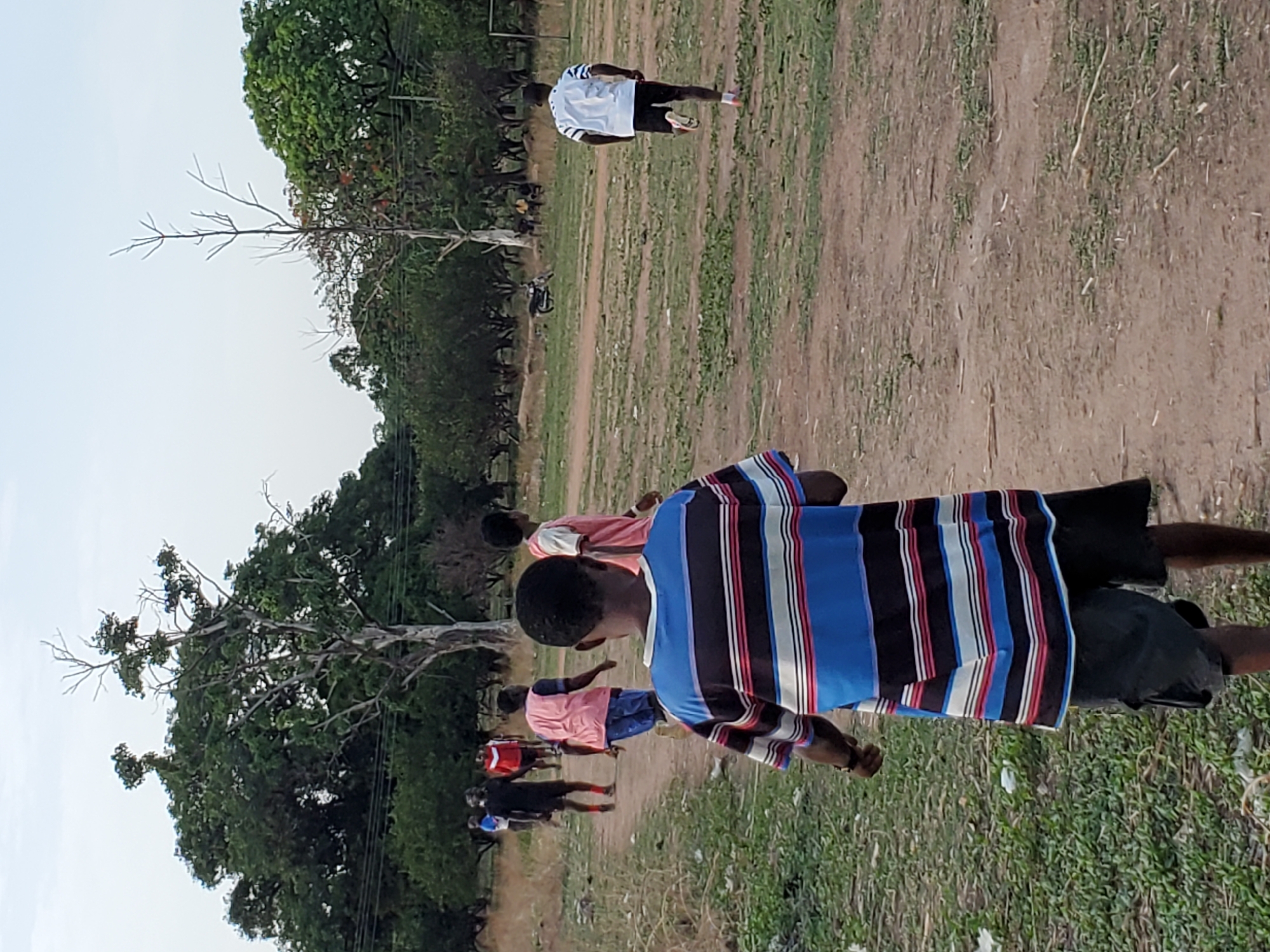 This media file captures an outdoor scene in Mararaba, Nigeria, featuring several young people in a dry, unpaved field under a bright sky. The ground is predominantly light brown earth with scattered patches of sparse green grass. Numerous trees, some with rich green foliage and others with bare branches, line the edges of the field, suggesting a natural or undeveloped area.
In the foreground, a young person is seen from behind, wearing a distinctive long-sleeved shirt with vertical stripes of blue, black, white, and red, paired with dark shorts. Further back, another individual is visible in a light pink or peach shirt and dark shorts. Towards the far left of the frame, a person in a white shirt with black stripes and dark shorts walks away from the camera. Several other figures, dressed in various casual attire including red and dark clothing, are dispersed across the mid-ground and background, bringing the total visible individuals to at least six.
A simple goalpost structure, possibly made of metal, is partially visible in the far background, indicating that the field might be used for sports, likely soccer. The people appear to be casually gathered, standing, or walking, suggesting an informal sporting event or outdoor play session. The clear, bright sky suggests it is daytime, possibly late afternoon given the soft light. No text is visible in the image.