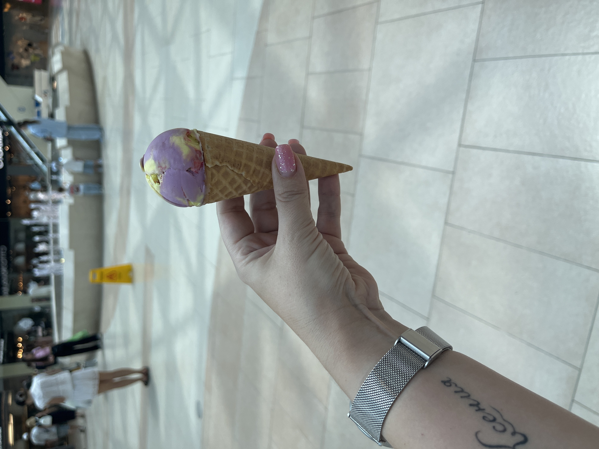 The image shows a close-up of a person's hand holding an ice cream cone with two scoops of ice cream, one purple and one yellow, in a waffle cone. The hand has a silver mesh watch on the wrist and a tattoo on the forearm that appears to be in Cyrillic script. The background is blurred, but it appears to be an indoor shopping mall with tiled flooring, fountains, and other shoppers. There is a yellow wet floor sign in the mid-ground. The overall impression is of a casual outing in a public space.
