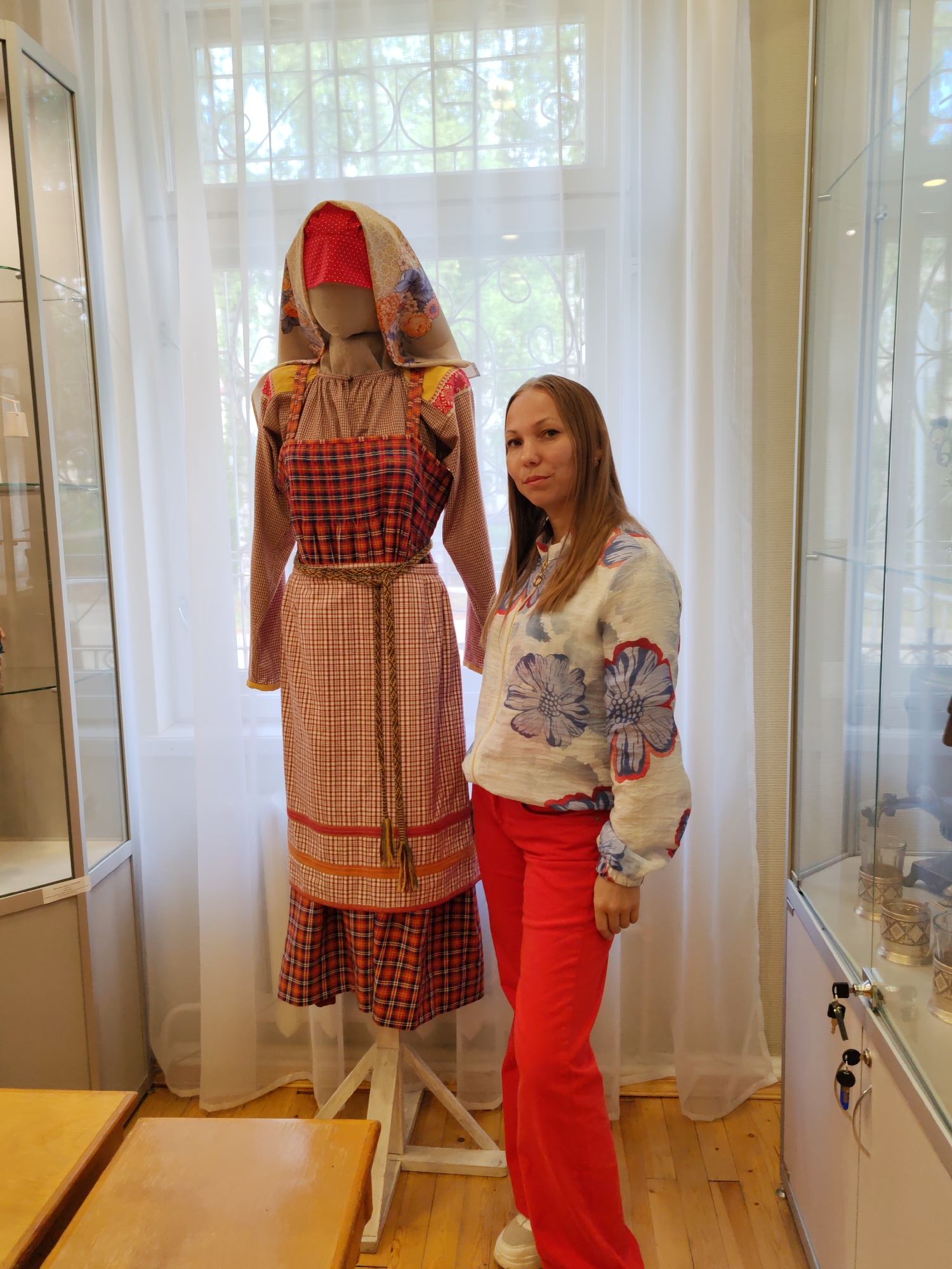 A young woman with long, brown hair stands to the right of a mannequin wearing traditional Russian folk attire in a museum setting. The mannequin displays a long-sleeved brown patterned shirt under a red and white checkered pinafore dress, complete with a woven belt and a colorful headscarf. The woman is dressed in a white blouse adorned with large blue and red floral prints and vibrant red pants. She is looking towards the camera with a neutral expression. The background features sheer white curtains, a window with visible greenery outside, and glass display cases on either side of the scene. The floor is made of polished wooden planks. The scene is indoors, likely a museum exhibition, and the objects displayed suggest a cultural or historical context. The presence of a mannequin dressed in traditional clothing and the display cases indicate an exhibit dedicated to local culture. Based on the metadata, the image was taken in Vizinga, Russia.