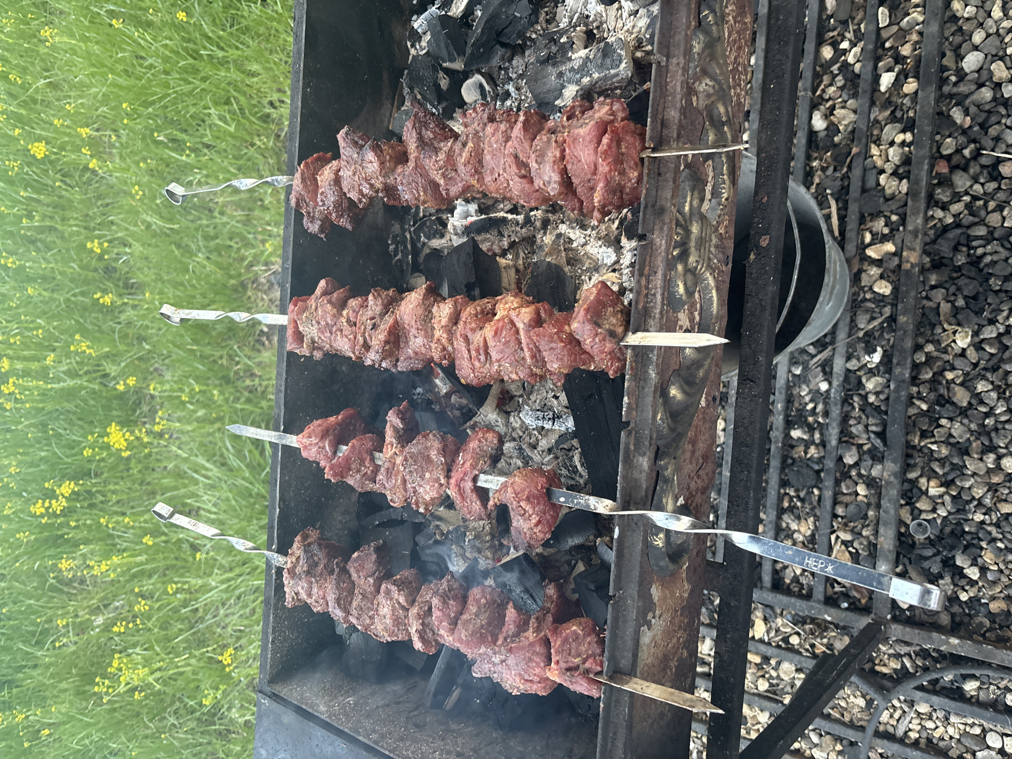 Four skewers of meat are cooking over charcoal on a grill. The location is impossible to determine solely from visual information, despite the possible "НЕРЖ" inscription on the skewer. The environment includes a gravel area and green grass with yellow flowers in the background.