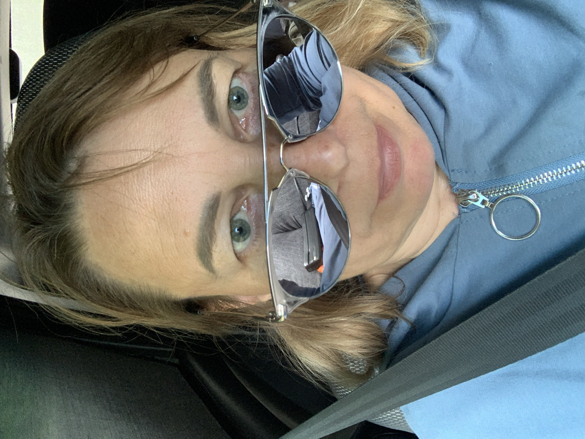 In a selfie taken from a frontal perspective, a woman with blonde hair and blue eyes is shown wearing sunglasses and a blue hooded jacket. The sunglasses are resting on her nose, obscuring her eyes, and the lenses reflect a gray interior, possibly of a vehicle. A black seatbelt is visible across her chest, suggesting she is a passenger or driver in a car. The background appears to be the interior of a car, with dark grey upholstery. The scene appears to be outdoors, as indicated by the natural lighting. There is no discernible indication of the specific location within Хаценчыцкі сельскі Савет, Belarus.