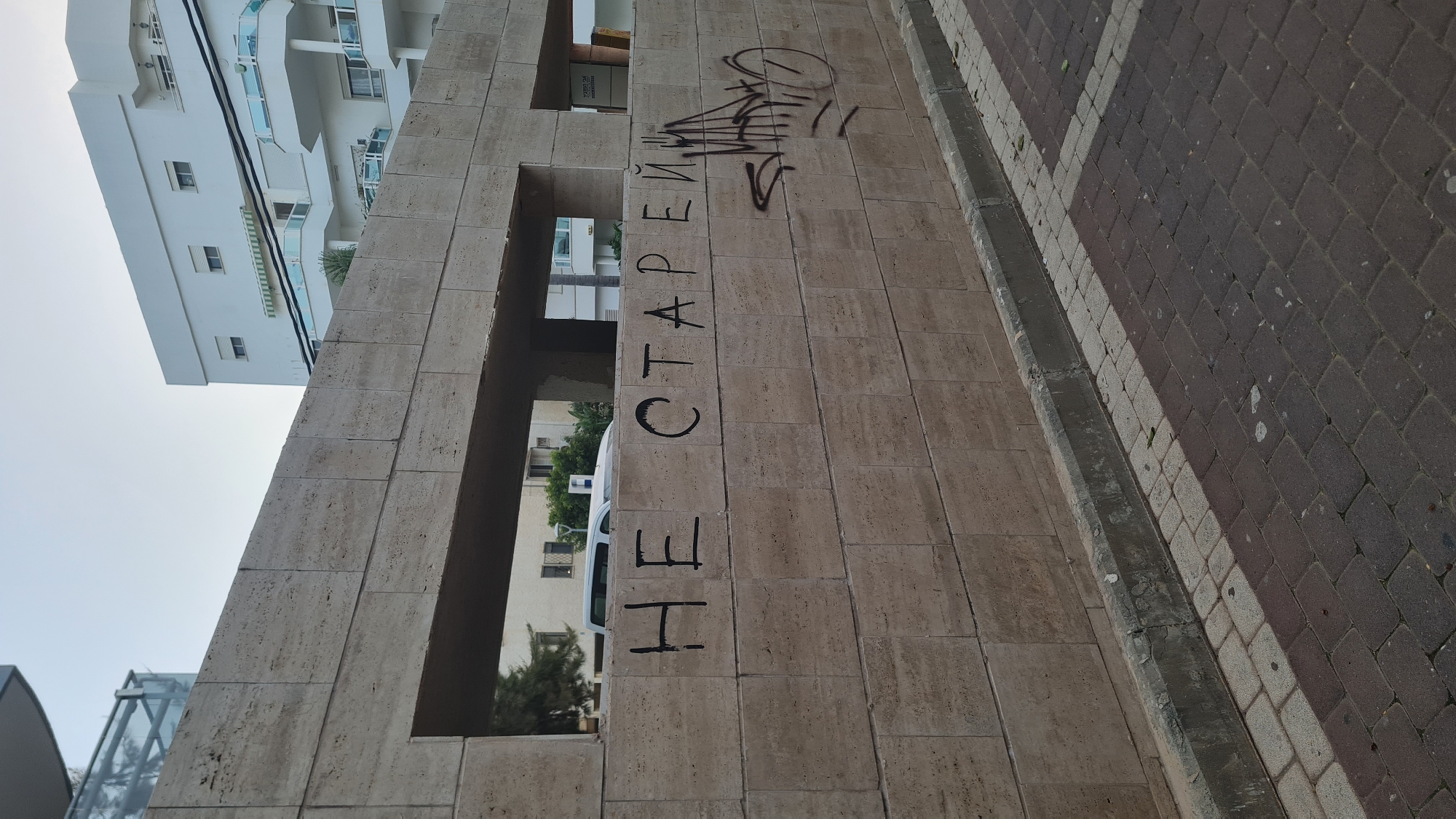 The image depicts a vertical view of graffiti in black spray paint on a light-colored stone structure, specifically the text "НЕ СТАРЕЙ" and some stylized graffiti. The text in Russian translates to "Don't get old." The structure appears to be a wall or pillar within an urban setting, likely a public space. A building with white facades and balconies is visible in the background. There is a pedestrian walkway paved with dark red bricks beside the stone structure, indicating this is an accessible public area. The image was taken in Nahariyya, Israel.
