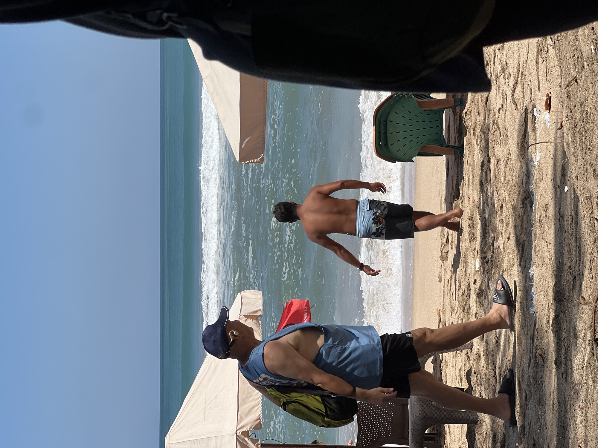 The image depicts a sunny day at Kuta Beach, Indonesia. In the foreground, a man wearing a blue sleeveless shirt, black shorts, a dark blue baseball cap, and black flip-flops walks from left to right, carrying a green backpack and a red object. He is wearing earbuds. Behind him, another man, shirtless and wearing patterned swim trunks, walks towards the ocean. The beach is sandy, with two cream-colored umbrellas providing shade. A green plastic chair is visible in the mid-ground. The ocean exhibits moderate waves with white foam, and the sky is clear and blue. The overall scene captures a moment of leisure at a popular Indonesian beach.