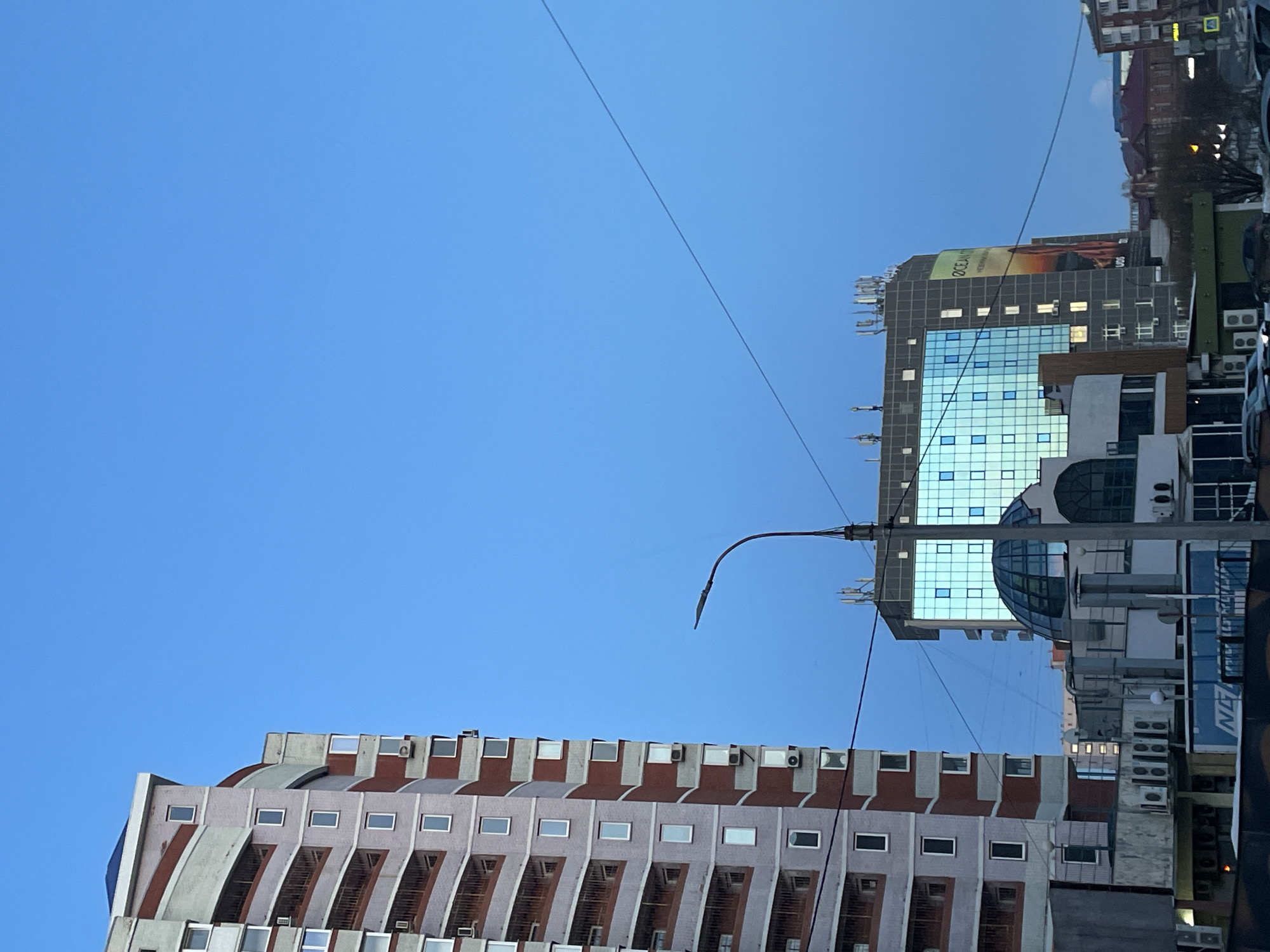 The image captures a street scene in Izhevsk, Russia. Dominating the view are two prominent buildings. The building in the lower left features a modern design with staggered, geometric balconies and horizontal lines of windows, each with an air conditioning unit. The other is a taller, glass-faced building with a rounded glass dome. A streetlight arches into the frame against a clear blue sky. Utility wires are visible overhead. Buildings and parked cars can be seen on the right.