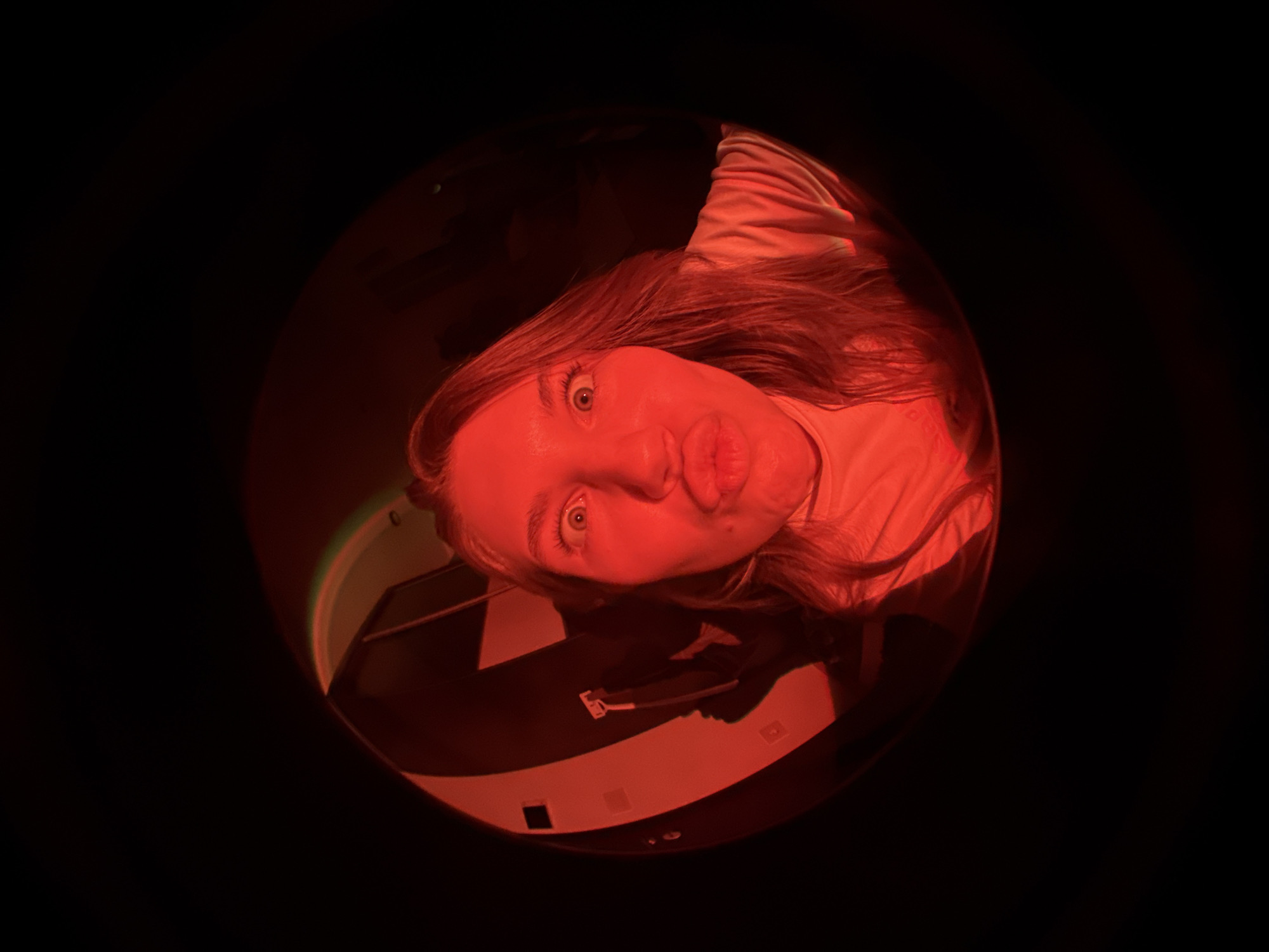 A fisheye lens captures a close-up view of a woman's face. The red light dominates the scene, casting a reddish hue on the woman's skin and hair. The woman is looking directly at the camera with a playful expression, pushing out her lips. In the lower portion of the circular image, part of a record player is visible. The rest of the scene is black. The image's location is unknown.