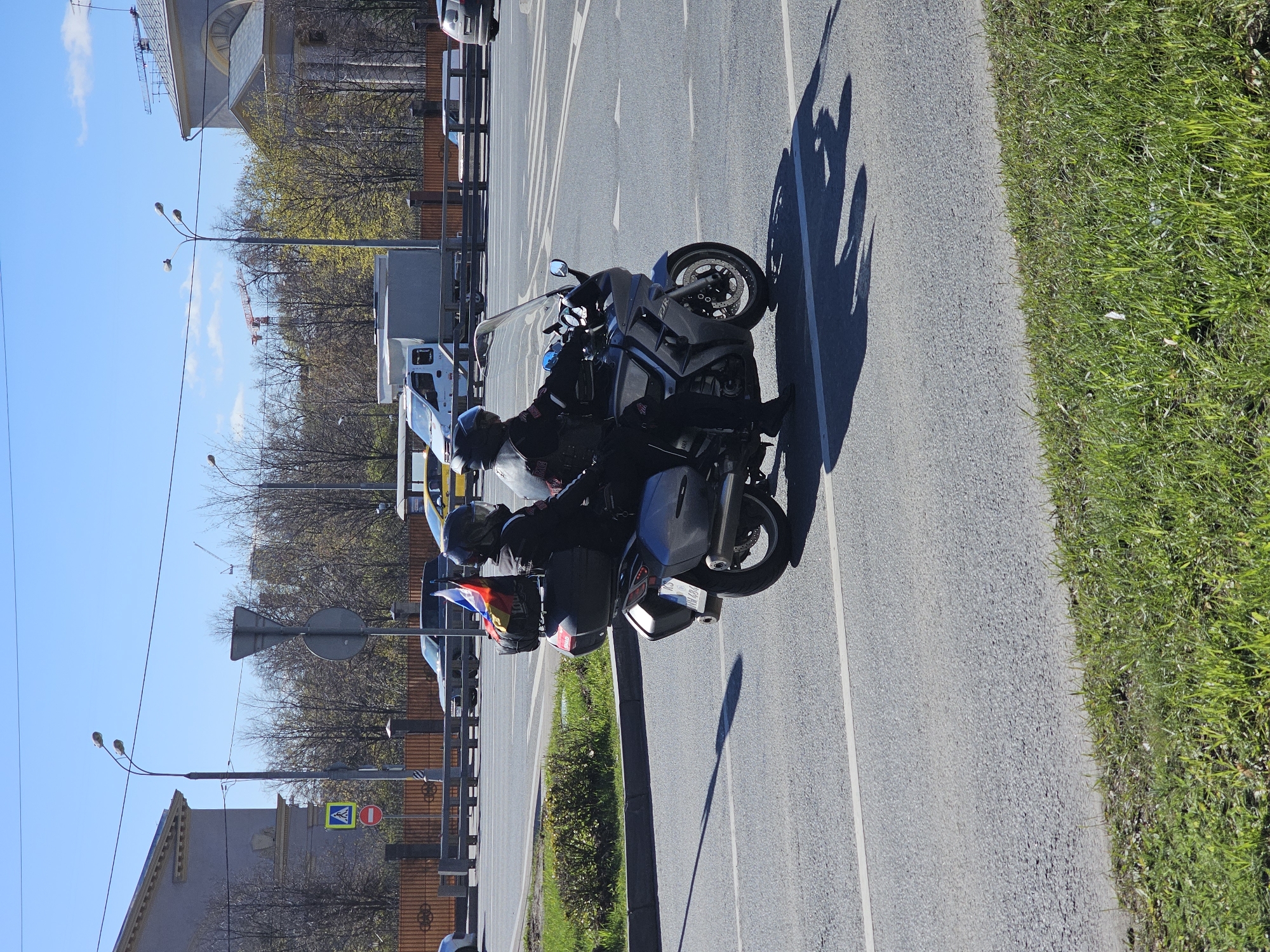 Two people on motorcycles are parked on the side of a road in Khimki, Russia. Both riders are wearing helmets and dark clothing. The motorcycle in the foreground has a Russian flag attached to the back. The road is paved with asphalt and has white lane markings. To the right of the road is a patch of green grass. On the other side of the road is a fence and buildings. The sky is blue with a few clouds. A construction crane can be seen in the distance.