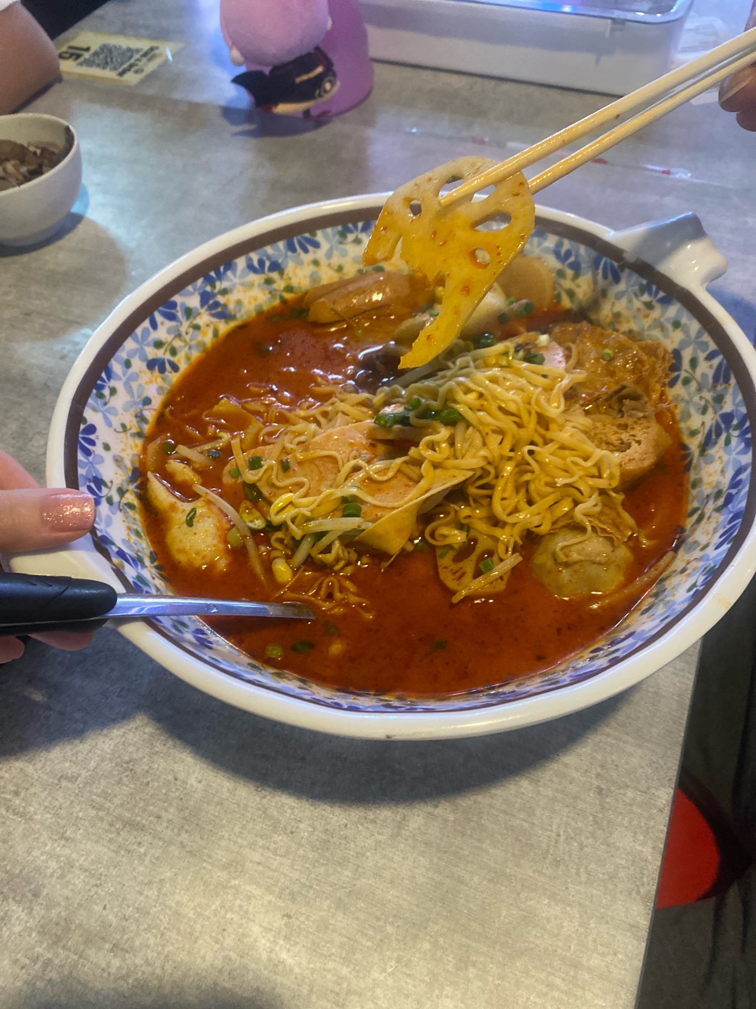 A person's hand, with a light pink, glittery fingernail, is holding chopsticks and lifting a slice of lotus root from a bowl of spicy soup. The lotus root is pale yellow with visible holes and appears to be seasoned with red chili flakes. The bowl is white with a blue floral pattern around the rim, and it contains noodles, bean sprouts, and other ingredients submerged in a rich, red broth. Another hand, visible on the left side of the image, is holding a black utensil, possibly a spoon, near the edge of the bowl. The table surface is gray and textured. In the background, a blurred purple plush toy with black eyes is visible on the table, and a QR code is present on a yellow card. The overall setting suggests a casual dining environment, possibly a restaurant in Kuala Lumpur, Malaysia, given the cuisine style and the mention of the city.