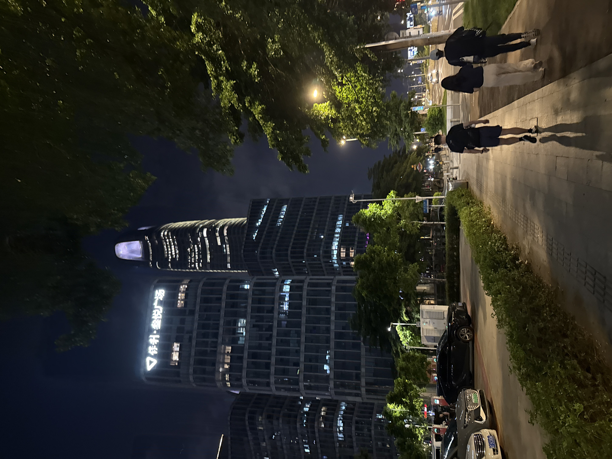 This media file captures a nighttime scene in Haizhu District, China, featuring a modern cityscape with illuminated buildings and a tree-lined pedestrian walkway. Three individuals are walking away from the camera on the walkway, casting long shadows. The first person appears to be wearing a black top and shorts, while the other two are dressed in dark clothing and light-colored pants.
Dominating the background are two tall, contemporary buildings with many windows, some of which are lit, indicating occupancy. The building on the left has a prominent triangular logo and white lettering that appears to be Chinese characters. The overall lighting suggests it is nighttime, possibly late evening, with artificial lights from the buildings, streetlights, and possibly car headlights illuminating the scene. The presence of trees suggests an urban park or a well-landscaped area. Two cars are parked on the side of the road adjacent to the walkway, one black and one grey, with visible license plates. The atmosphere appears calm and ordinary, depicting a typical evening in a developed urban area.