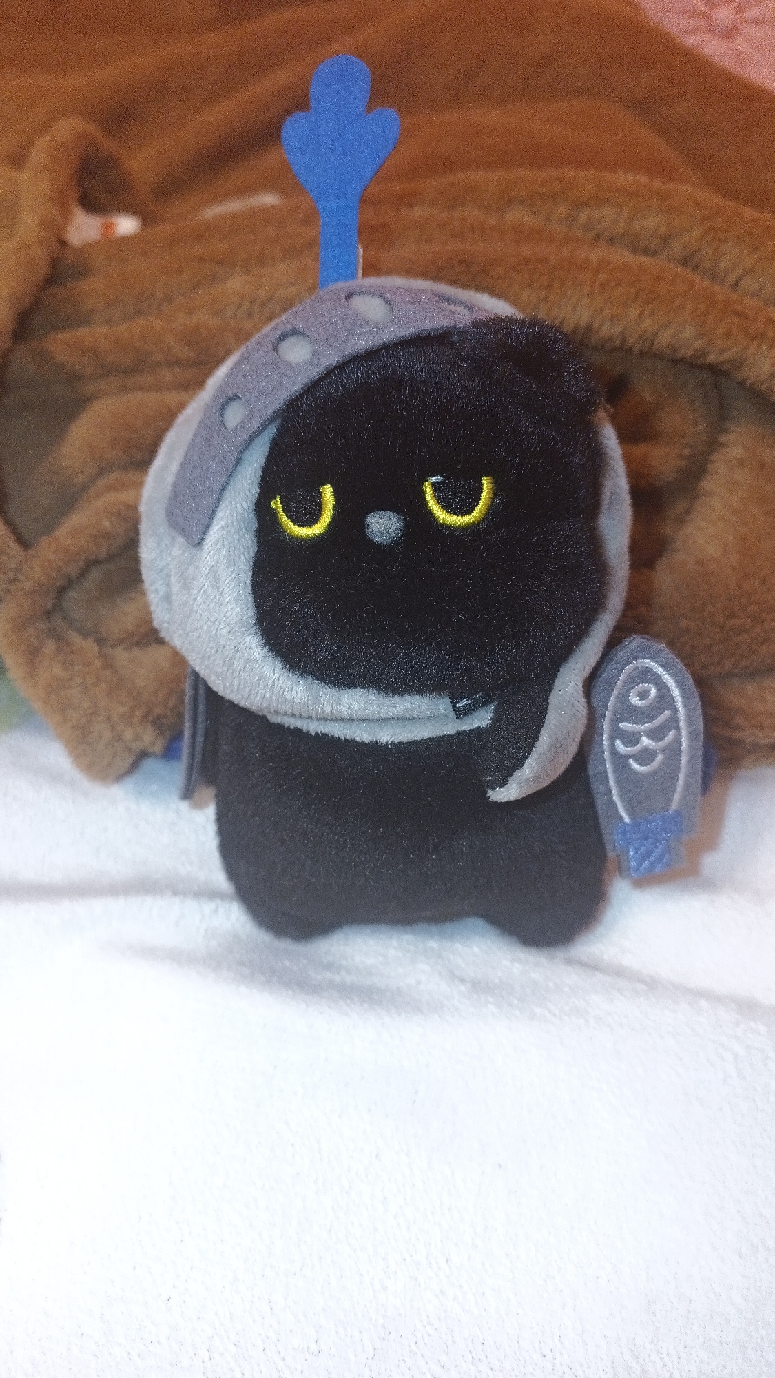 A plush toy of a black, rounded character is positioned on a white, textured surface, likely a blanket or cloth. The character has yellow crescent-shaped eyes and a small, dark nose. It wears a gray helmet with evenly spaced, circular indentations around the lower rim and a blue, stylized antenna extending upwards from the top. A gray, shield-like object with a blue, fish-shaped design is attached to its right side. The background consists of a blurred, brown fabric. The location in the city of Karnobat, Bulgaria is not determinable from the image.