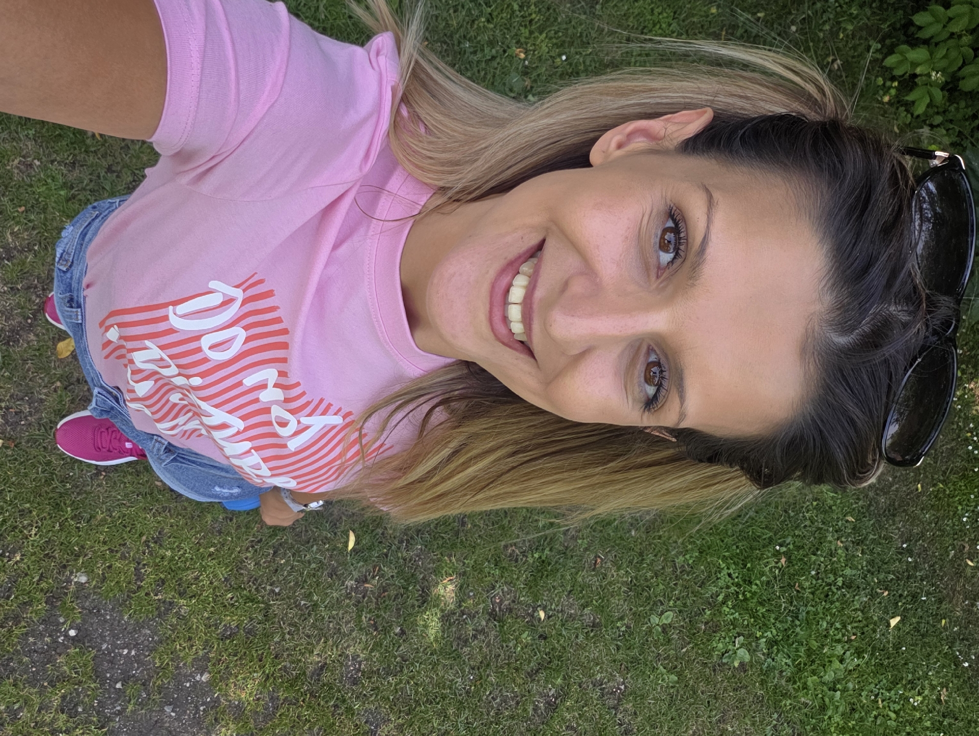 A woman is standing on a grassy area, likely a park or a backyard, with her sunglasses perched on her head. She is wearing a pink t-shirt with a graphic design and denim shorts. Her hair is pulled back in pigtails. The woman is smiling directly at the camera, and the photo appears to be a selfie. The background consists of green grass and some dappled sunlight, suggesting an outdoor setting during the day. No specific location in Munich, Germany can be identified from the image.