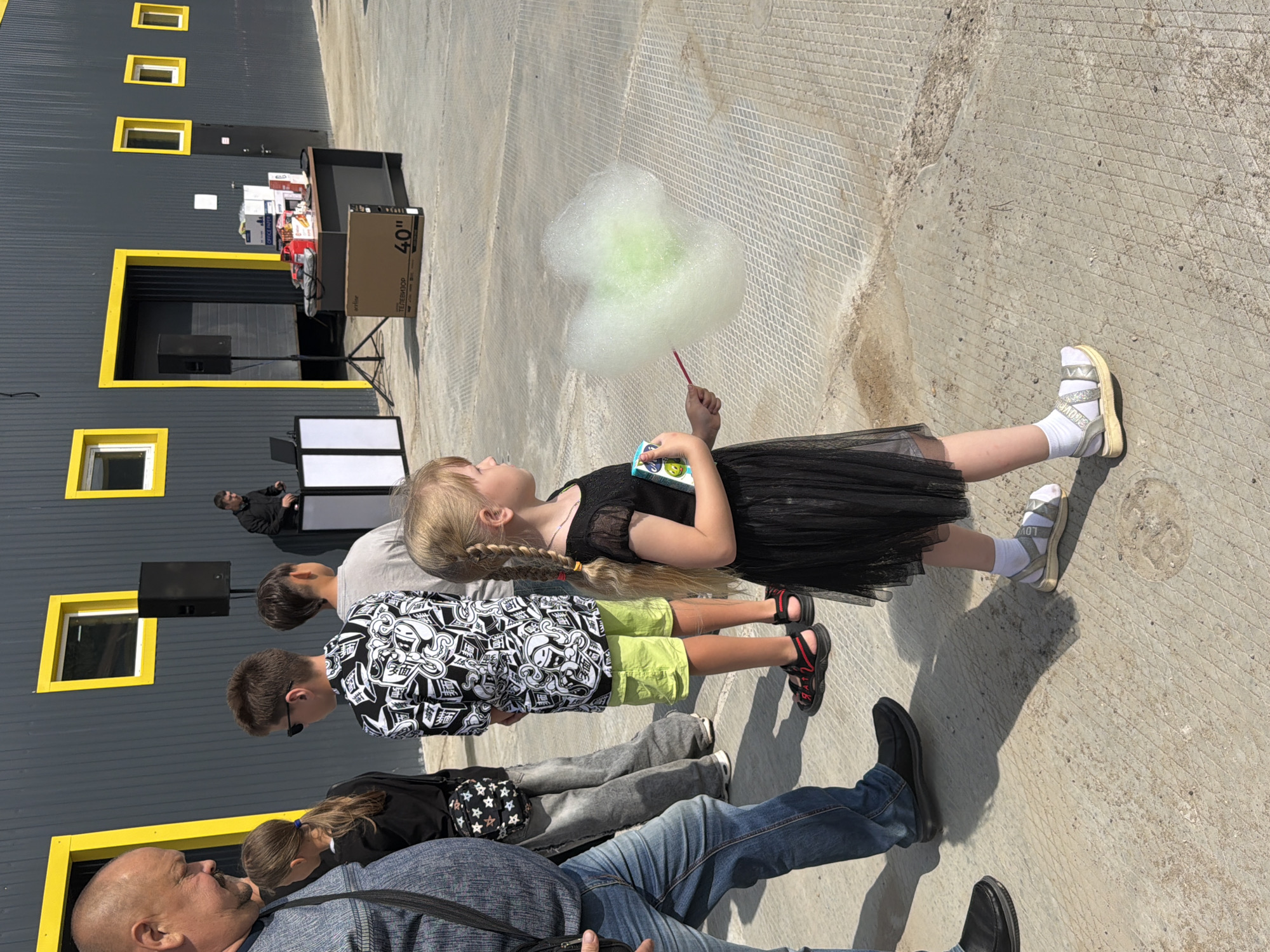 The image depicts an outdoor scene, likely in Chaikovsky, Russia, near a modern, grey building with bright yellow window frames and doorways. In the foreground, a young girl with blonde braided hair, wearing a black dress and sandals, holds a stick with a large cloud of green and white cotton candy. To her left, a man, a girl, and two boys are partially visible, facing away from the camera. The man is wearing a blue denim shirt and jeans, the girl in the background is wearing a black top and a patterned crossbody bag, and the boys are in casual attire. Further back, there is a DJ setup with two white columns and a laptop, with a person operating it. A table displays several boxes and items, and a 40-inch television box is on the ground in front of it. The ground is a paved surface with a diamond pattern. The lighting suggests it is daytime with bright sunlight.