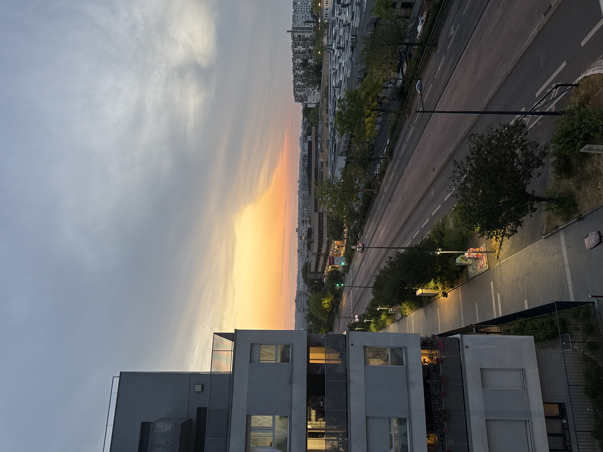 The image depicts an urban scene at dusk, with a large, modern apartment building dominating the left side. The building has multiple floors with balconies and windows, some of which are illuminated, indicating occupancy. A paved walkway with streetlights runs along the side of the building, bordered by greenery. Adjacent to the walkway is a multi-lane road, appearing relatively empty, with a few distant vehicles visible. The background features a skyline of low-rise and high-rise buildings, suggesting a densely populated urban area. The sky exhibits the colors of sunset, with a gradient of orange and yellow near the horizon, transitioning to gray and cloudy tones overhead. The overall impression is a serene, twilight view of a residential and commercial district.