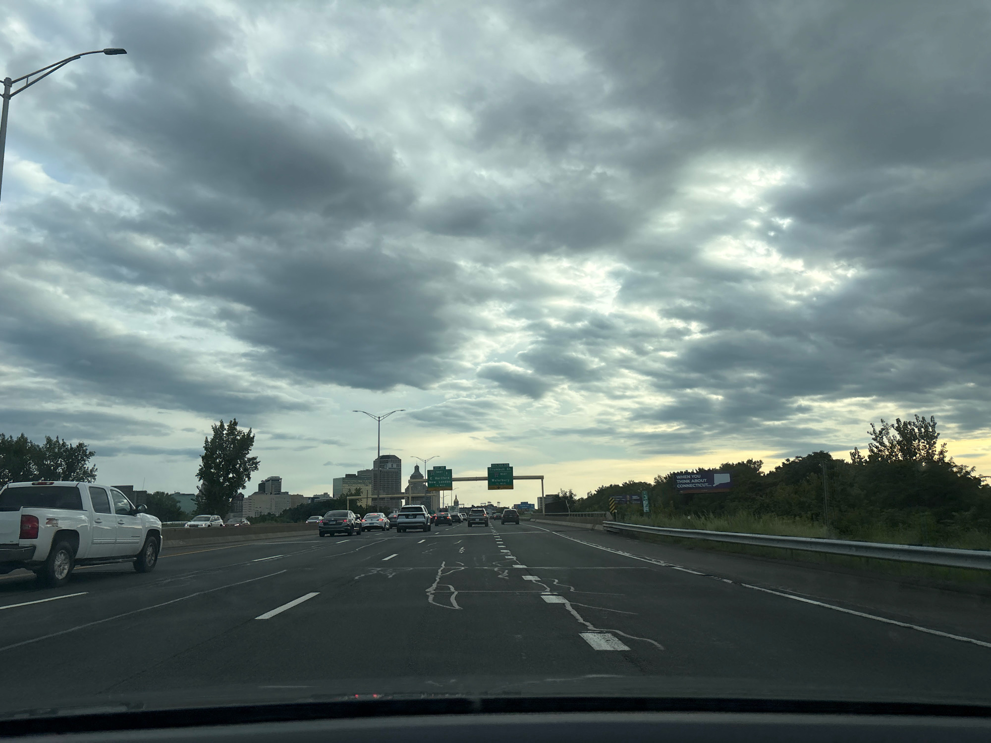The image depicts a view from a vehicle traveling on a multi-lane highway, likely in or near Hartford, Connecticut. The sky is overcast with dramatic, heavy grey clouds. On the right side of the highway, a billboard with text that appears to say "WHEN YOU THINK ABOUT CONNECTICUT..." is visible. The highway is lined with trees and vegetation on both sides, with a metal guardrail separating the lanes from the shoulder. In the distance, the skyline of a city, consistent with Hartford, is visible, featuring several buildings. Several vehicles are traveling on the highway in the same direction as the viewpoint. On the left side of the highway, a white pickup truck is prominently visible in the foreground. Overhead streetlights are also present. The road surface shows some white markings and what appear to be faded or repaired lane lines.