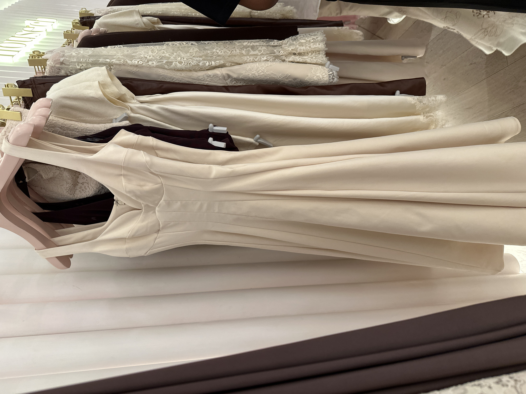 The image shows a close-up view of several dresses hanging on a rack in what appears to be a retail setting. The dresses are in shades of white, cream, and brown, and some have lace detailing. They are arranged on hangers of different colors, including gold and pink. The background is blurred and suggests a shop interior. There is no information to suggest a London location.