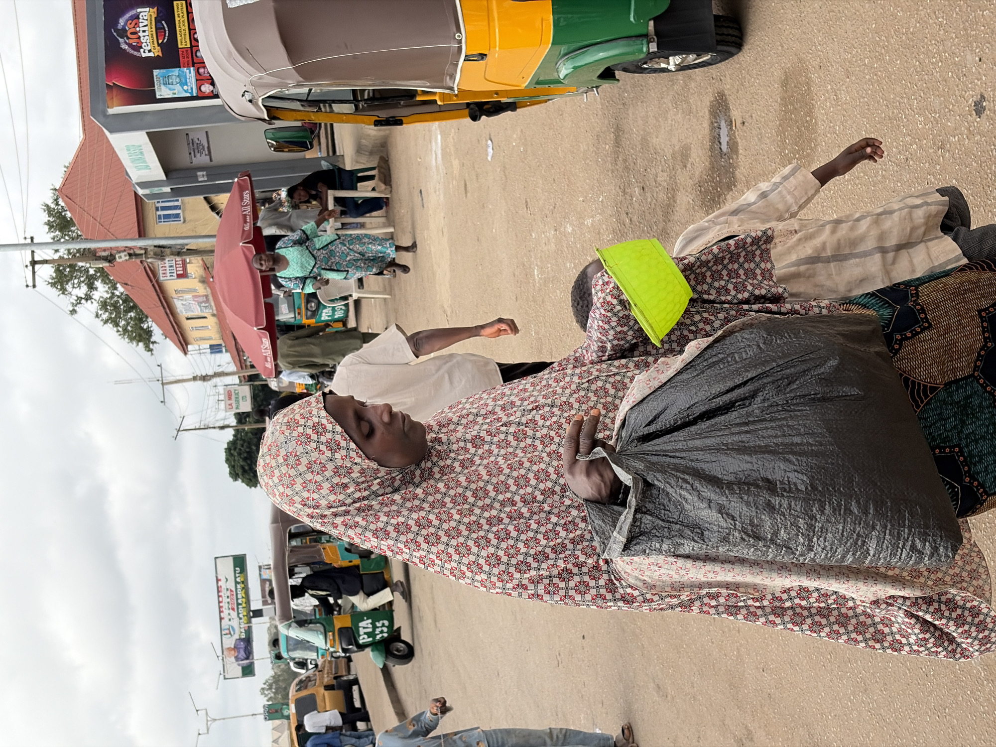 This media file captures a bustling street scene in Gura Top, Nigeria, on an overcast day. The main subject in the foreground is a woman, seen from the chest up, wearing a white, red, and black patterned hijab and dress, carrying a large, dark grey plastic bag over her shoulder. Her expression is calm. Behind her, another person is partially visible, wearing a beige striped garment and carrying a bright neon yellow bag on their head.
The scene is populated by numerous individuals engaged in daily activities. In the mid-ground, people are seen standing and sitting on plastic chairs under large red market umbrellas. Further back, several green and yellow auto-rickshaws (tuk-tuks) are visible, some parked, others possibly in motion. Buildings line the street, featuring various signs and architectural details. Power lines crisscross the upper part of the image against the cloudy sky.
Visible text on signs and billboards includes: "PTA-135", "LA MED PHARMACY LTD", "SHADGA PREMIO BREAK", "we are All Stars", and a large advertisement for "JOS Festival OF PRAISE WORSHIP" with location details "32TH PHILADELPHIA, PO TINGI JOS PLATEAU". The overall impression is that of a lively urban or commercial area.