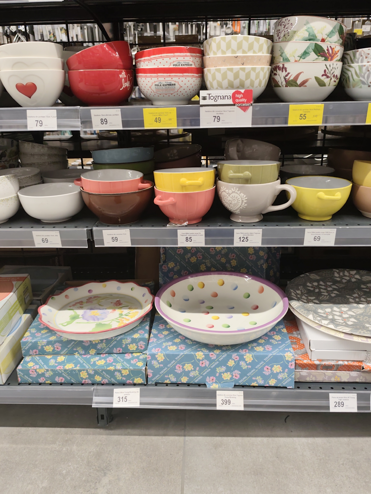 A wide-angle, eye-level shot captures shelves of ceramic bowls and plates in a retail setting. The lighting is even, and the focus is sharp across the scene. The shelves are organized with rows of various tableware items, primarily bowls of different sizes, colors, and patterns. On the top shelf, a stack of white and red bowls is visible, some with heart designs and text like "with love" and "POLE EXPRESS". Next to them are stacks of bowls with geometric and floral patterns.
The second shelf displays a mix of single bowls, including white, red, and yellow options, and a large white mug with a heart design. Below this, the third shelf holds more bowls in various colors like brown, pink, and green, along with a white and gray bowl.
The bottom shelves are dominated by decorative serving dishes and plates. One prominent item is a large, round serving dish with a scalloped edge, adorned with colorful polka dots and floral designs. This dish is placed on top of stacked boxes with a floral pattern. To the right of this, several stacked plates with intricate floral patterns are visible.
Price tags are attached to the shelves, indicating prices in the local currency (likely Moldovan Leu, given the location context). Some of these tags include product names such as "Salatiera din portelan", "Casa", and "Farfurie".
The overall environment appears to be a well-lit retail store, likely a home goods or kitchenware section. The flooring is a light gray, concrete-like material. No people are visible in the image. The context provided suggests this scene is in Chișinău, Moldova, implying the store is a local establishment there.