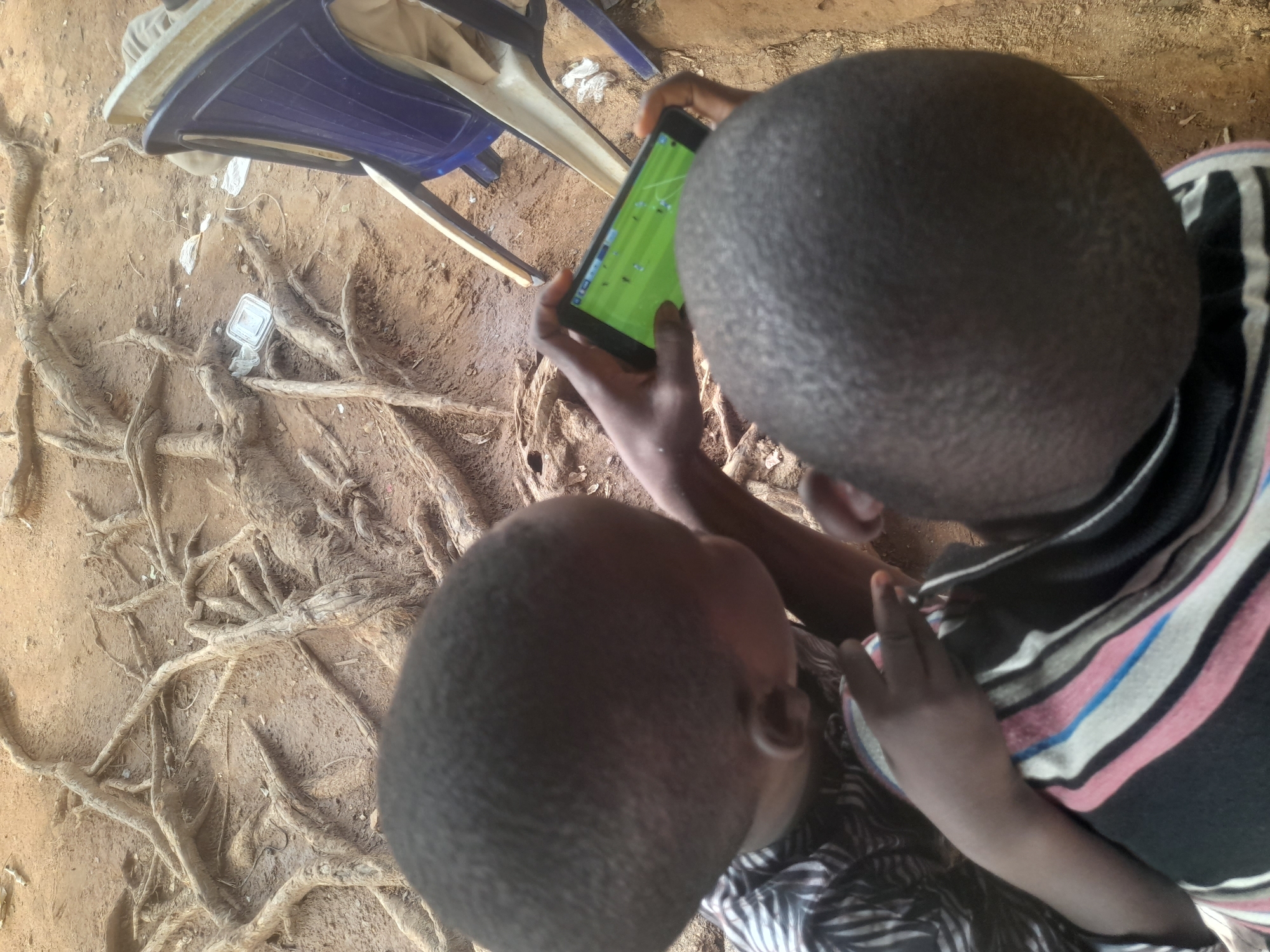 Two children are focused on a mobile device displaying a soccer game. They sit near the exposed roots of a tree in a dirt yard. A blue plastic chair sits nearby. The location is in Akwanga, Nigeria.