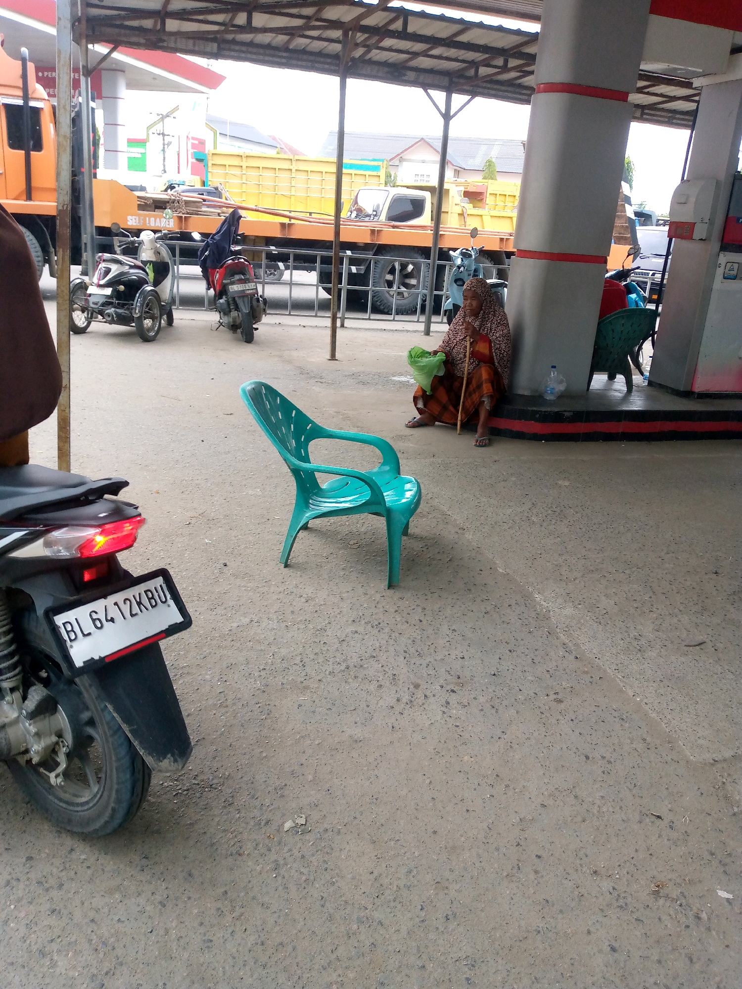 The image depicts a scene at a gas station in Keudebayu, Indonesia. In the foreground, the rear of a motorcycle with a license plate reading "BL 6412KBU" is visible, its red taillight illuminated. To the right, a bright teal plastic chair stands on the concrete ground. Further back, a woman wearing a patterned headscarf and a checkered skirt sits on the ground leaning against a pillar, holding a light green bag and a wooden stick. Behind her, a green plastic chair is visible. In the background, several motorcycles are parked. A yellow truck with a flatbed is also present, along with a part of an orange truck on the left. The gas station canopy and pillars are prominent elements of the environment.