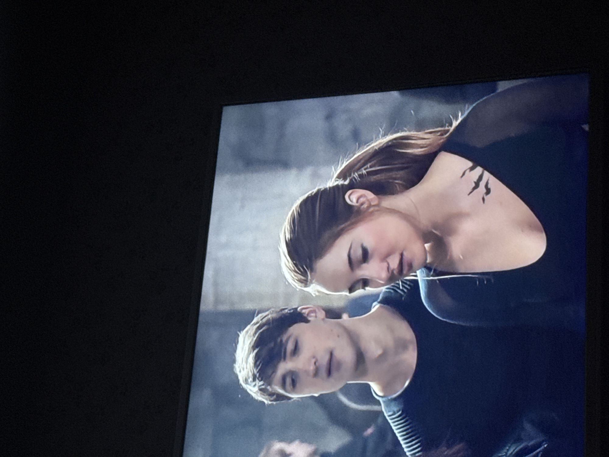The image is a photograph of a television screen displaying a scene of two individuals, a man and a woman. The woman is positioned with her head near the man's neck. She has light skin, light brown hair pulled back, and a tattoo of birds near her collarbone. She wears a dark top. The man has short, dark hair and wears a dark striped shirt. The background appears to be a gray, possibly concrete, setting. The image is a close-up shot from what seems like a television display. This image does not contain information about Russia or Mytishchi.