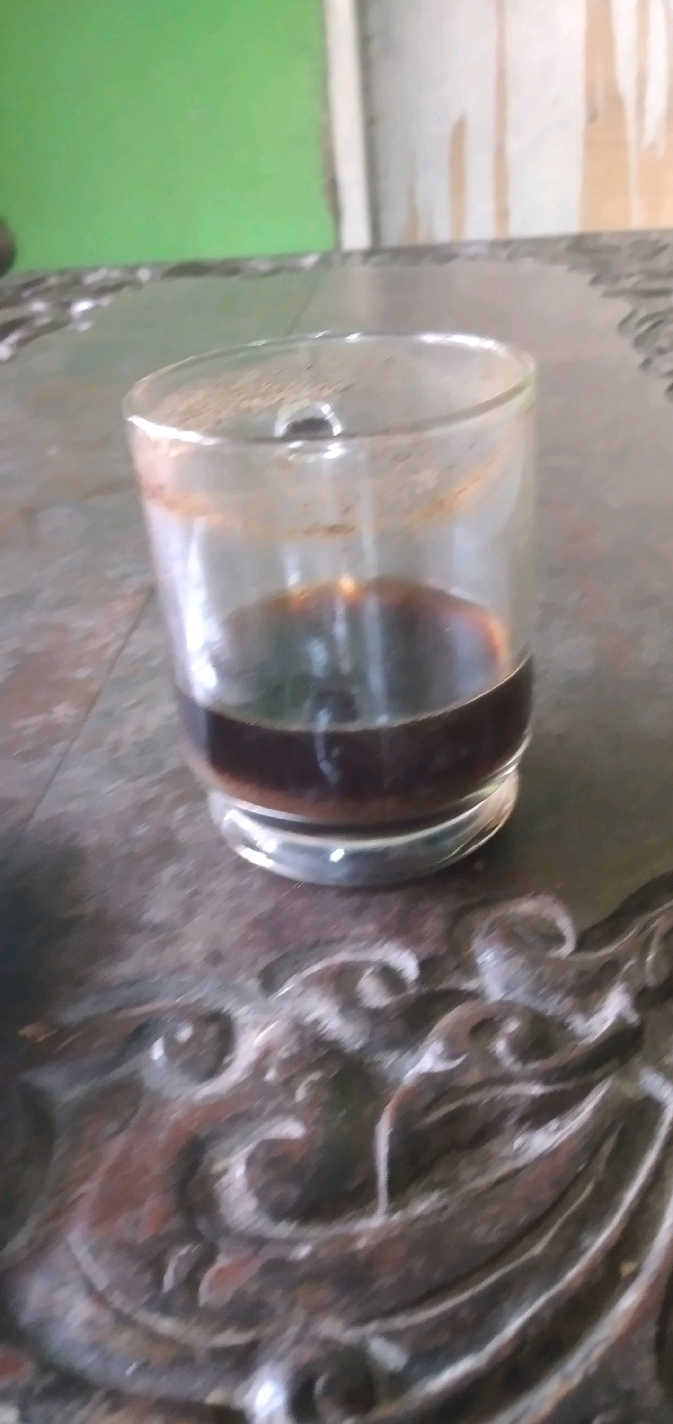 A shot of a glass of coffee on a textured table. The table has intricate carvings of what appears to be a face or animal. The background is blurry, but there is a green wall.