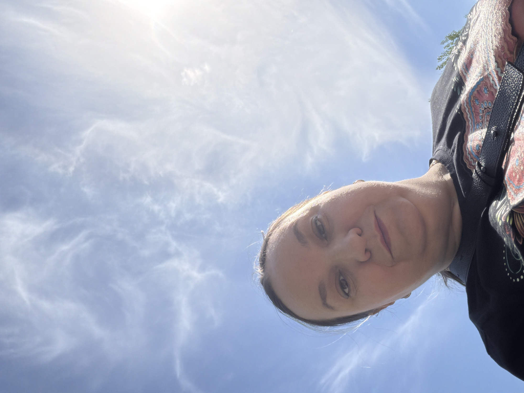 The image shows a woman looking down at the camera, positioned below her. The background is a bright blue sky with wispy, cirrus clouds. The sun is shining brightly, creating a glare in the upper left corner. She is wearing a dark-colored top, and a patterned scarf or shawl is visible at her right shoulder. A black bag strap crosses her body. The photo's location within the city of Chaikovsky, Russia cannot be determined from the visual information provided.