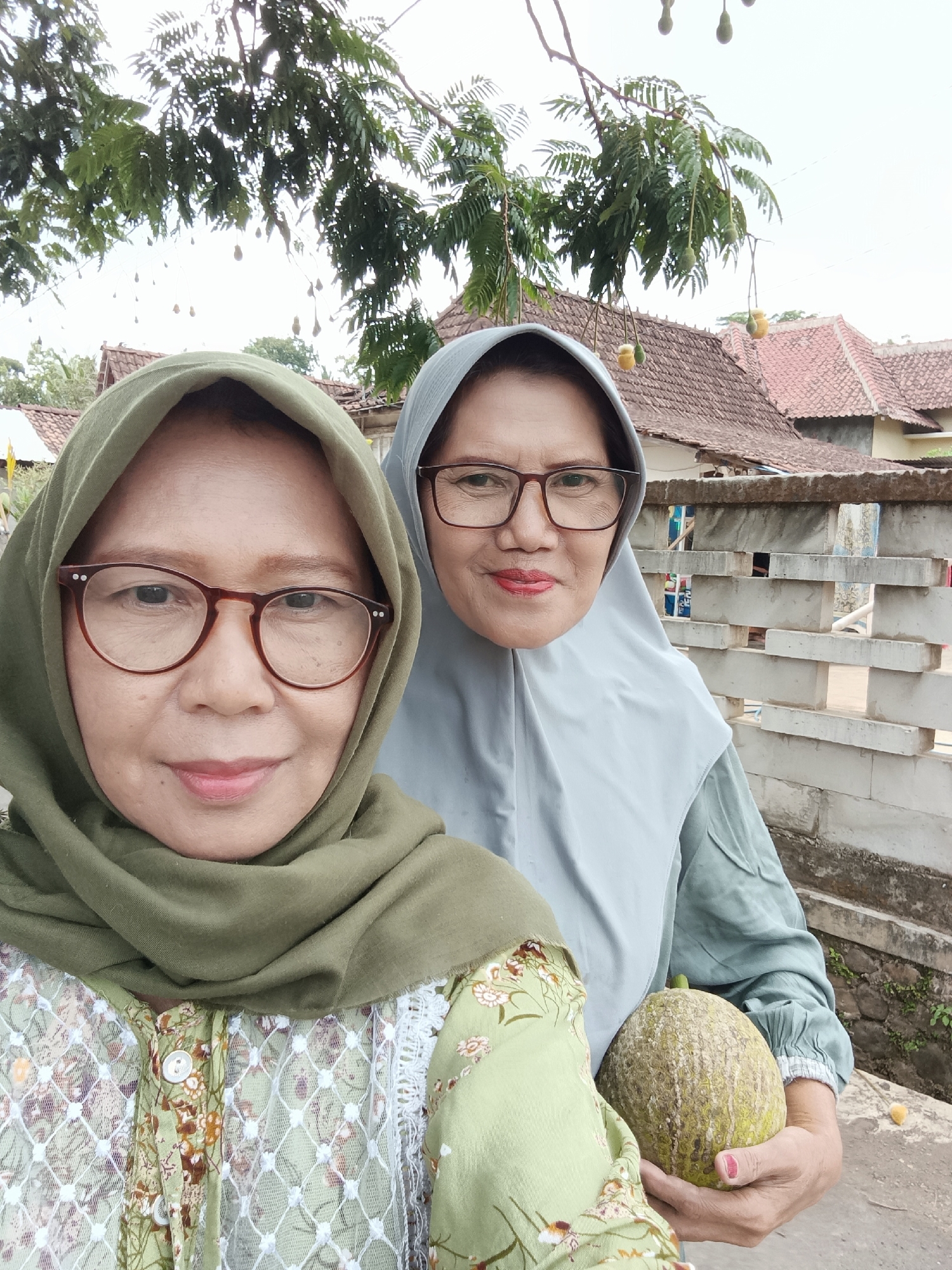 Two adult women, likely Indonesian, are taking a selfie outdoors. The woman in the foreground wears a green hijab and a floral patterned top. She has glasses with dark frames and is smiling. The woman behind her wears a light blue hijab and a matching colored shirt. She also wears glasses and has red lipstick on. She is holding a large, rough-skinned fruit. In the background, there are traditional Indonesian houses with tiled roofs, a stone fence, and a tree with hanging fruit. The scene appears to be in a rural or suburban area in Tayu, Indonesia.